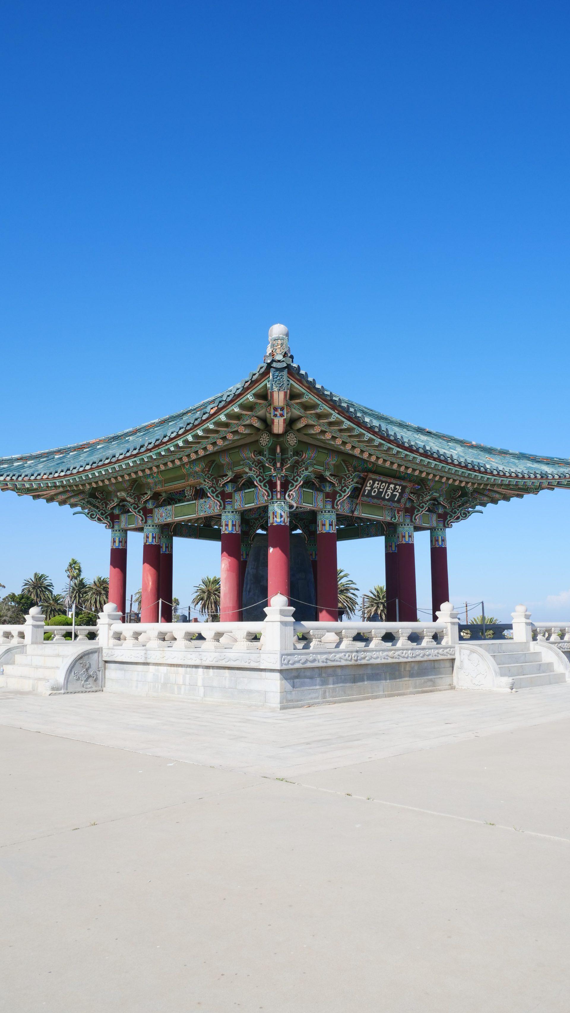 A traditional Korean-style pavilion with ornate green and red details stands under a clear blue sky, surrounded by palm trees and a white stone platform.