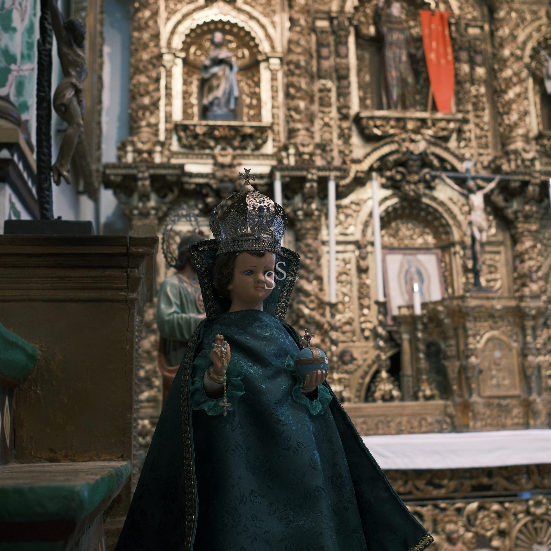A small statue of a crowned child dressed in ornate green robes stands in front of an intricately decorated gold altar inside a church.