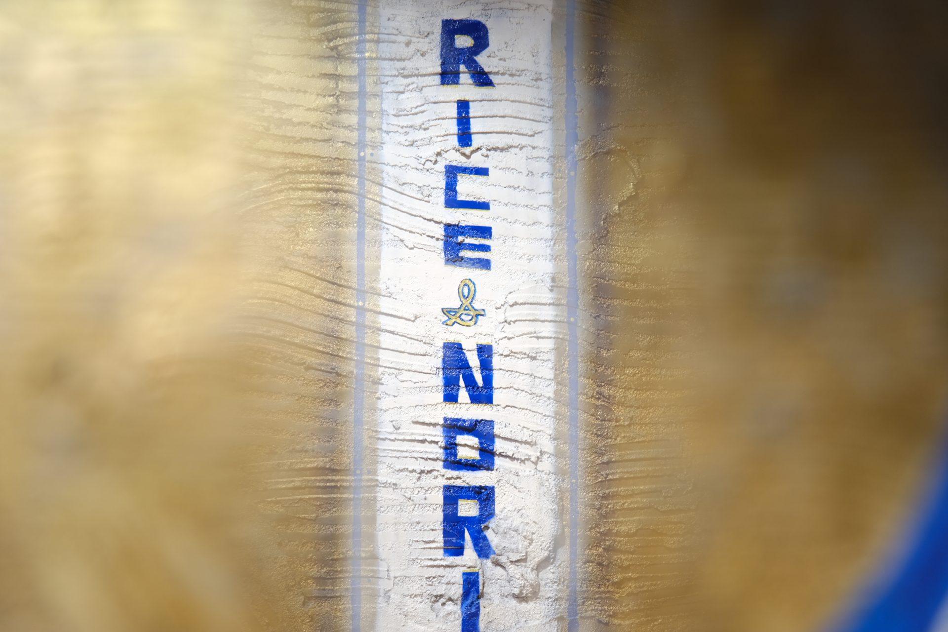 A close-up of a wall with the words RICE NORI written vertically in blue letters on a white strip, surrounded by a golden-brown textured background.