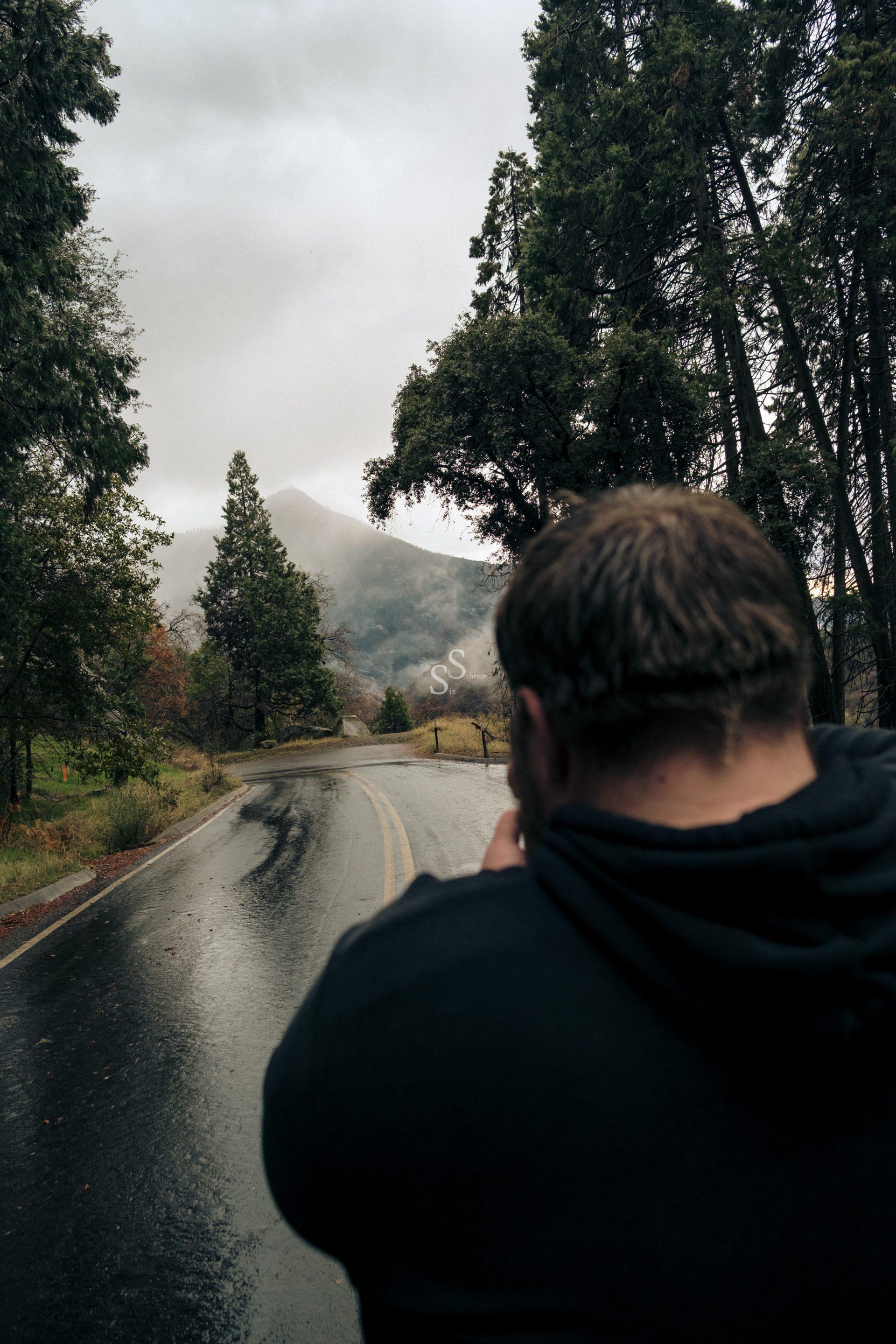 A person in a dark hoodie stands on a wet, curving road surrounded by tall trees, looking toward misty mountains under a cloudy sky.