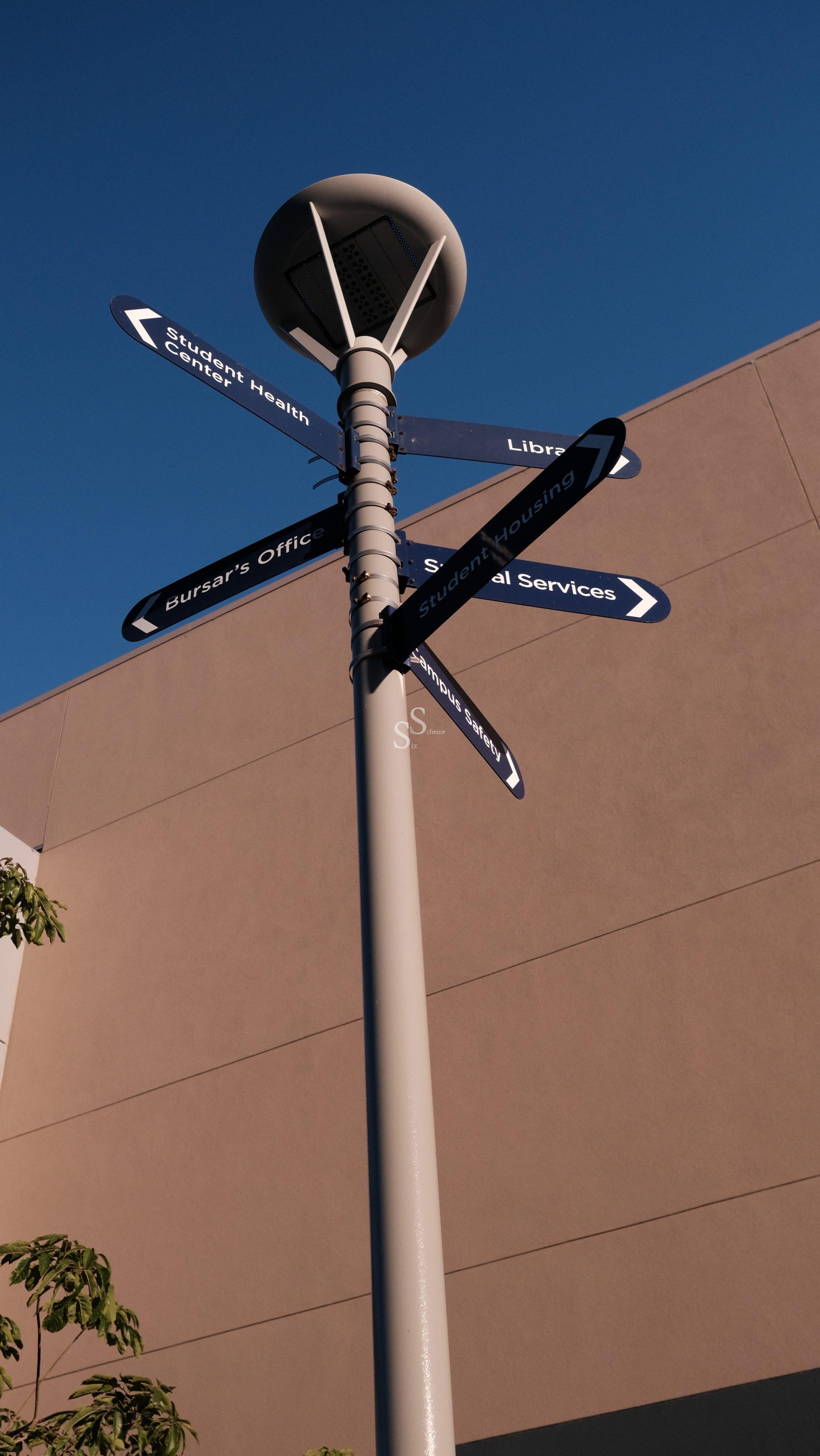 A tall signpost with multiple blue directional signs points to locations like Student Health Center, Library, Bursars Office, Student Housing, and Student Services, set against a beige building and clear blue sky.