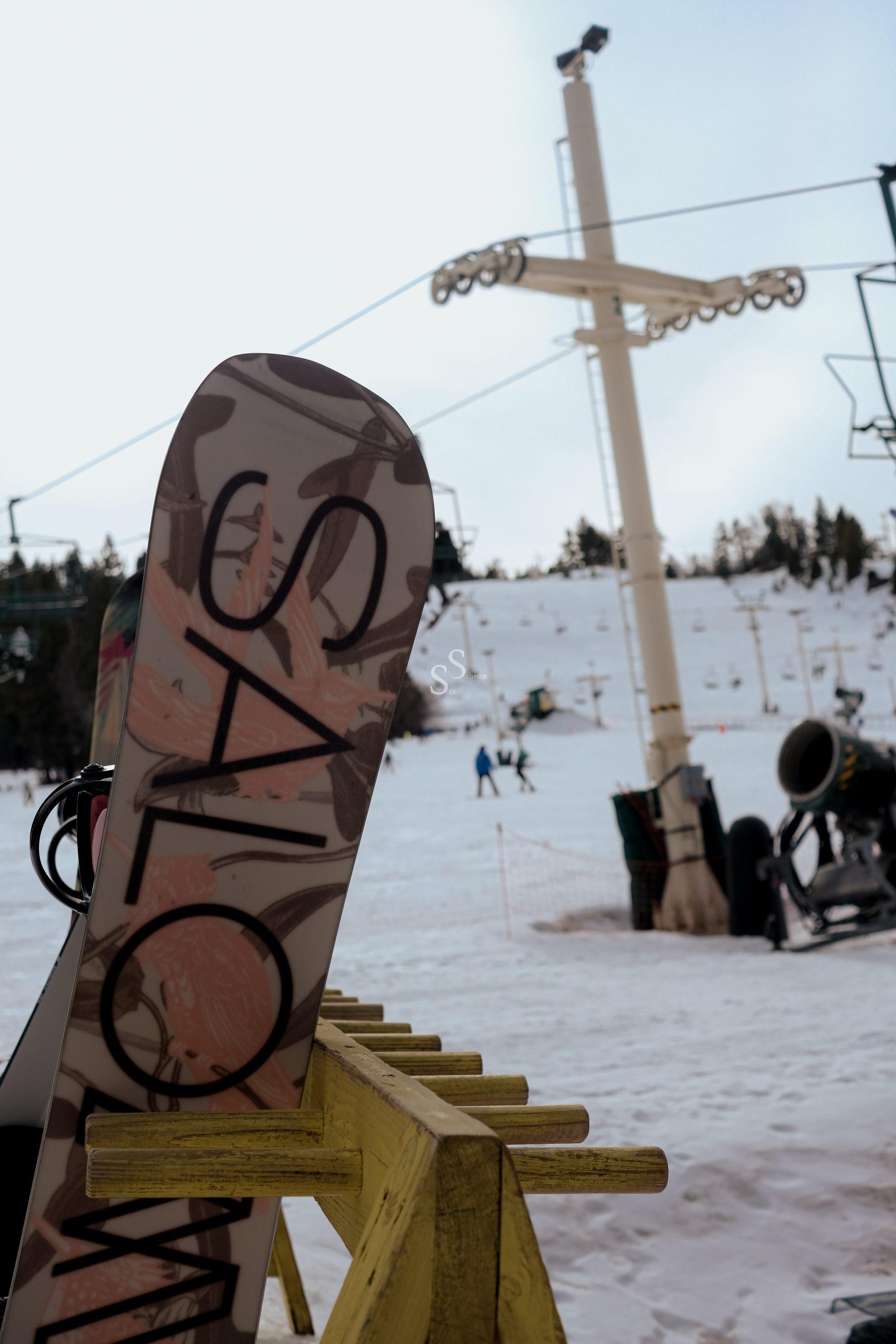 A close-up of a snowboard with SALOMON written on it, resting on a yellow rack near a snowy ski slope with a ski lift and people in the background.