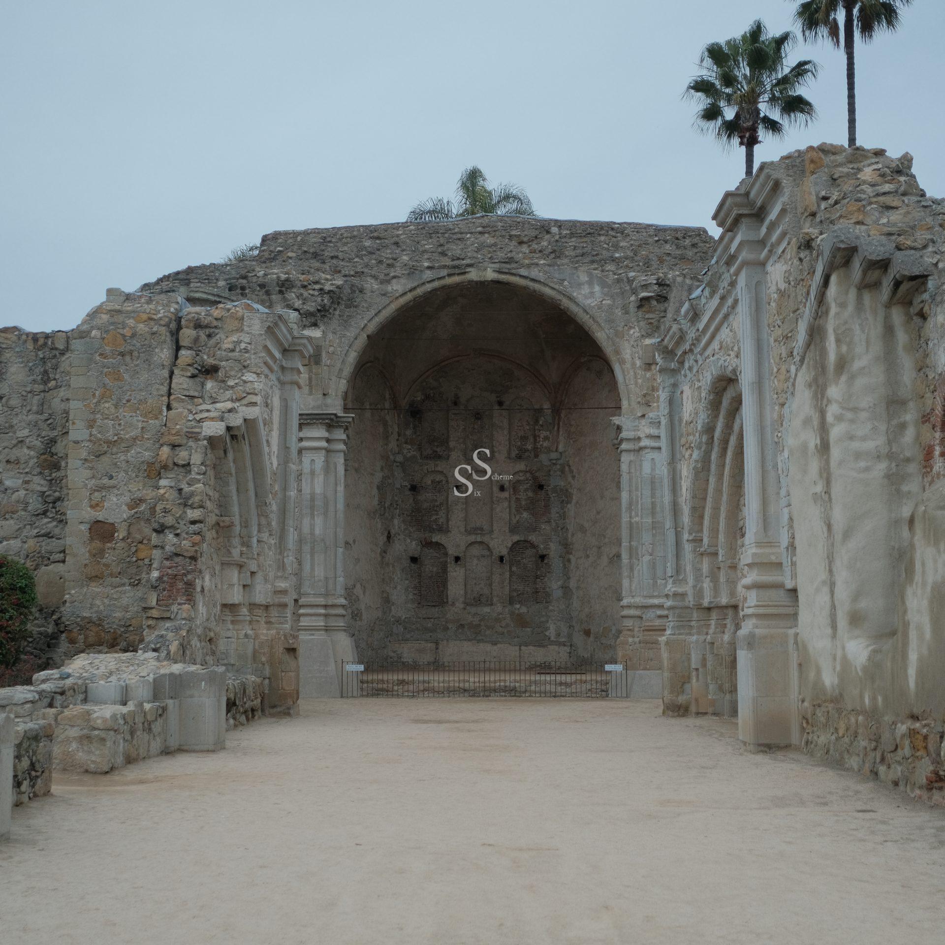 Ruins of a historic stone building with arched entryways, weathered walls, and partial columns. Two palm trees rise above the structure, and the sky is overcast. The ground is sandy and the ruins appear ancient.