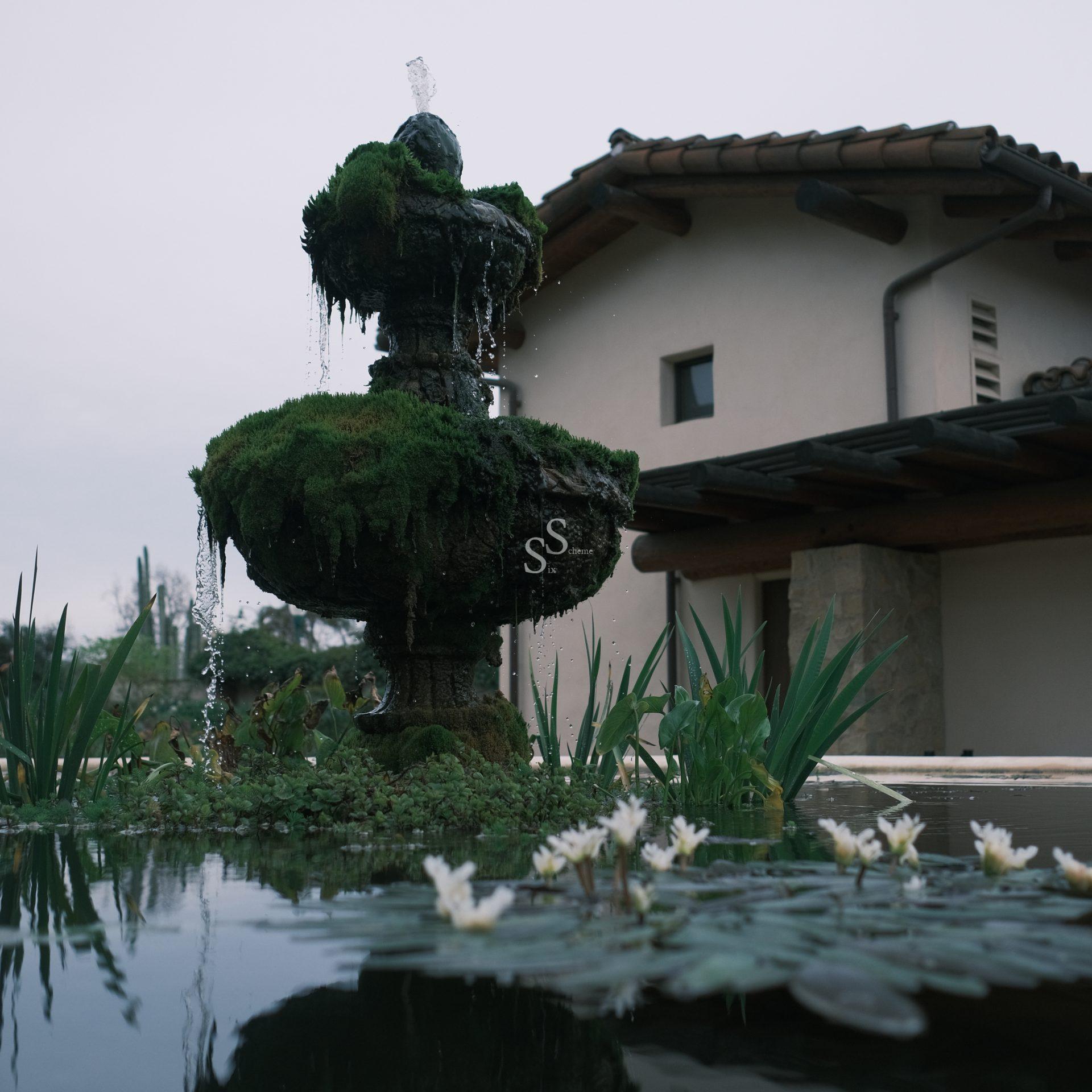A moss-covered stone fountain with water trickling down, surrounded by a pond with white water lilies, is set in front of a rustic house under an overcast sky.