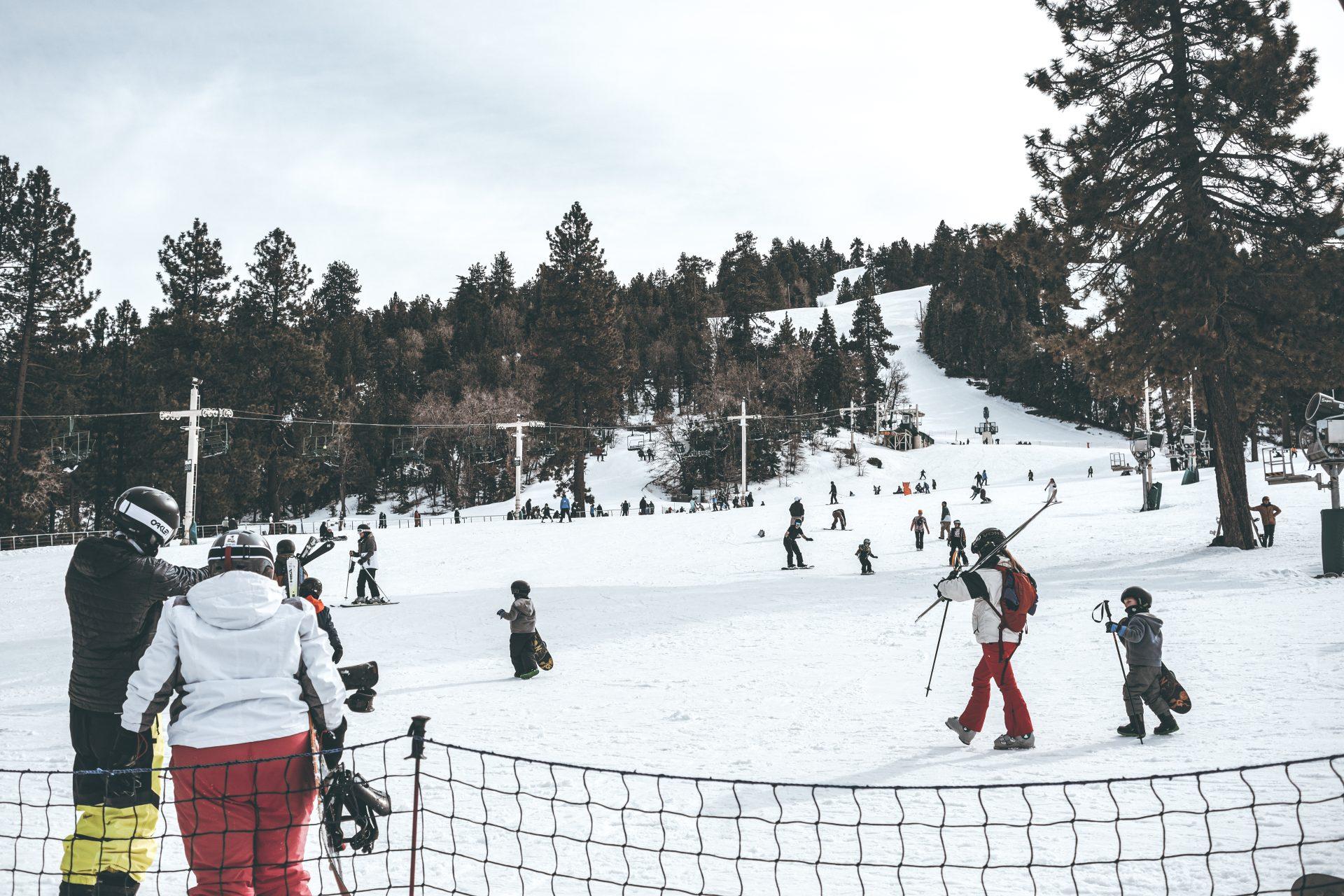 People dressed in winter gear are skiing and snowboarding on a snowy slope surrounded by trees, with a ski lift and more skiers in the background under a cloudy sky.