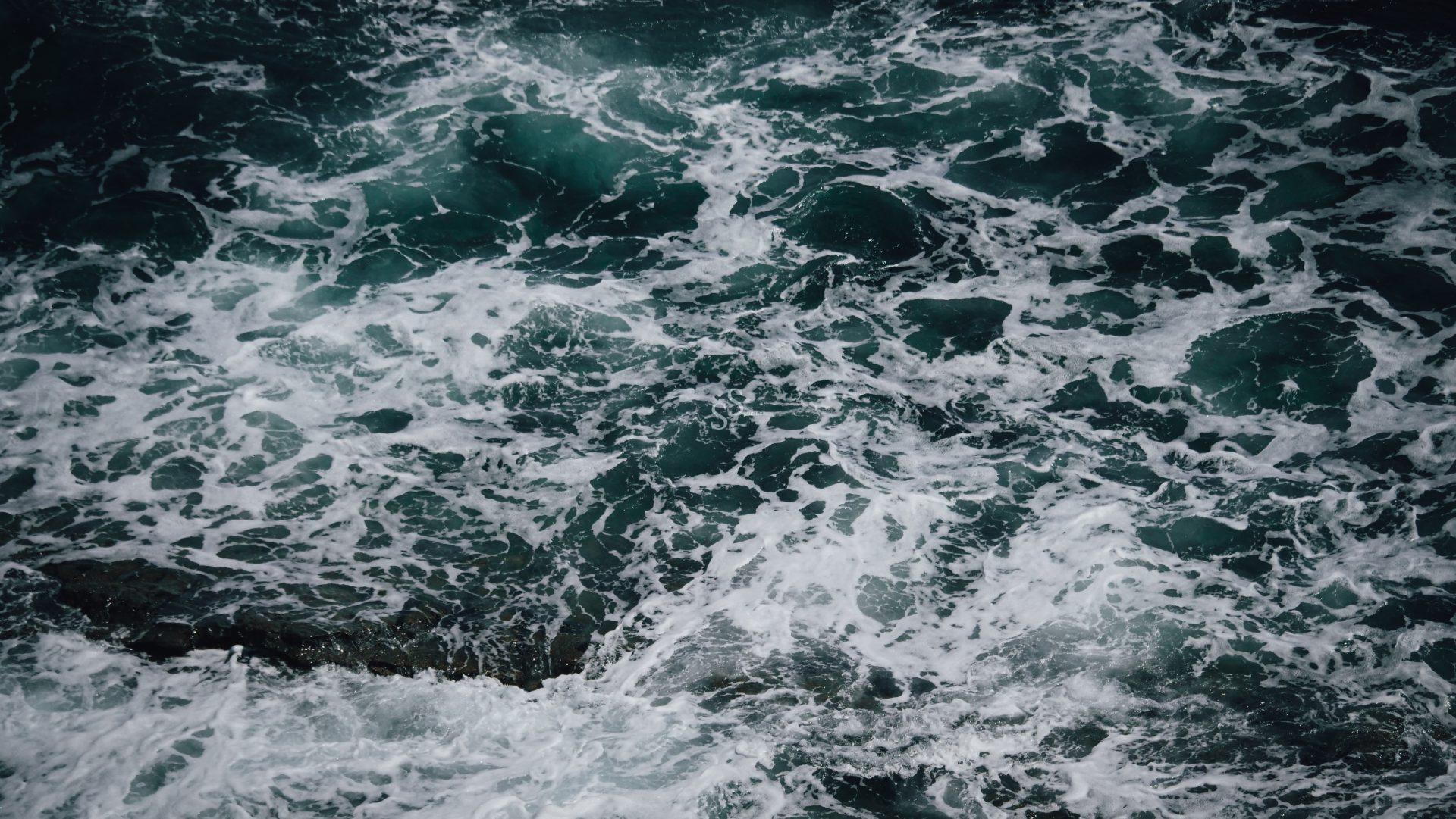 Waves of dark teal ocean water churn with white foam and bubbles, creating a swirling, textured surface pattern.