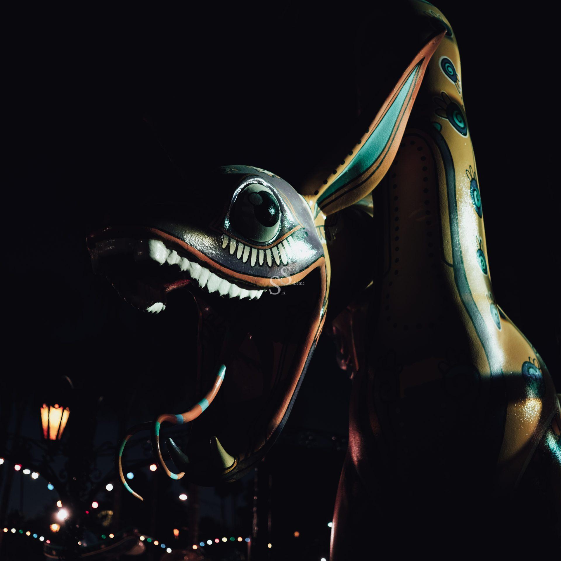 A colorful, large alebrije sculpture resembling a fantastical animal with big eyes and an open mouth, illuminated against a dark background with festive string lights and a streetlamp nearby.