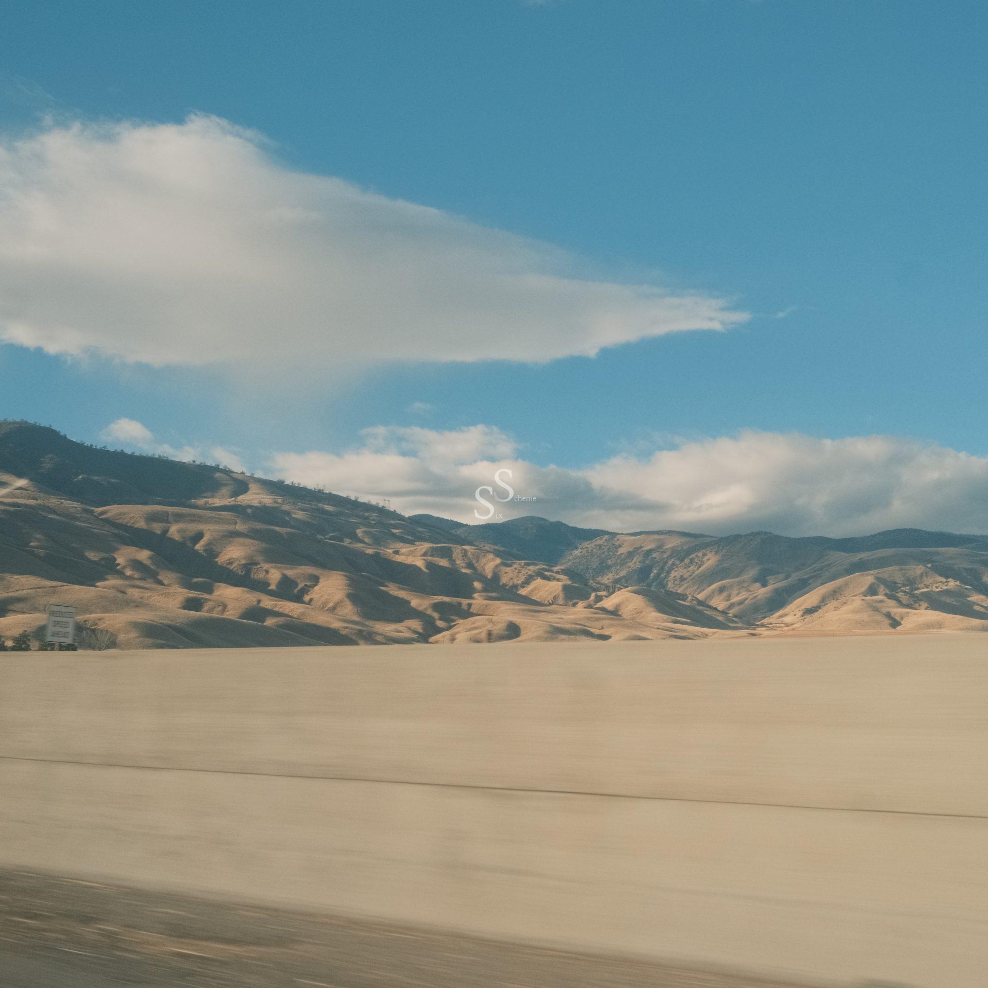 A concrete highway runs alongside rolling, dry hills under a clear blue sky with scattered clouds. The scene suggests a warm, sunny day in a rural or semi-arid landscape.