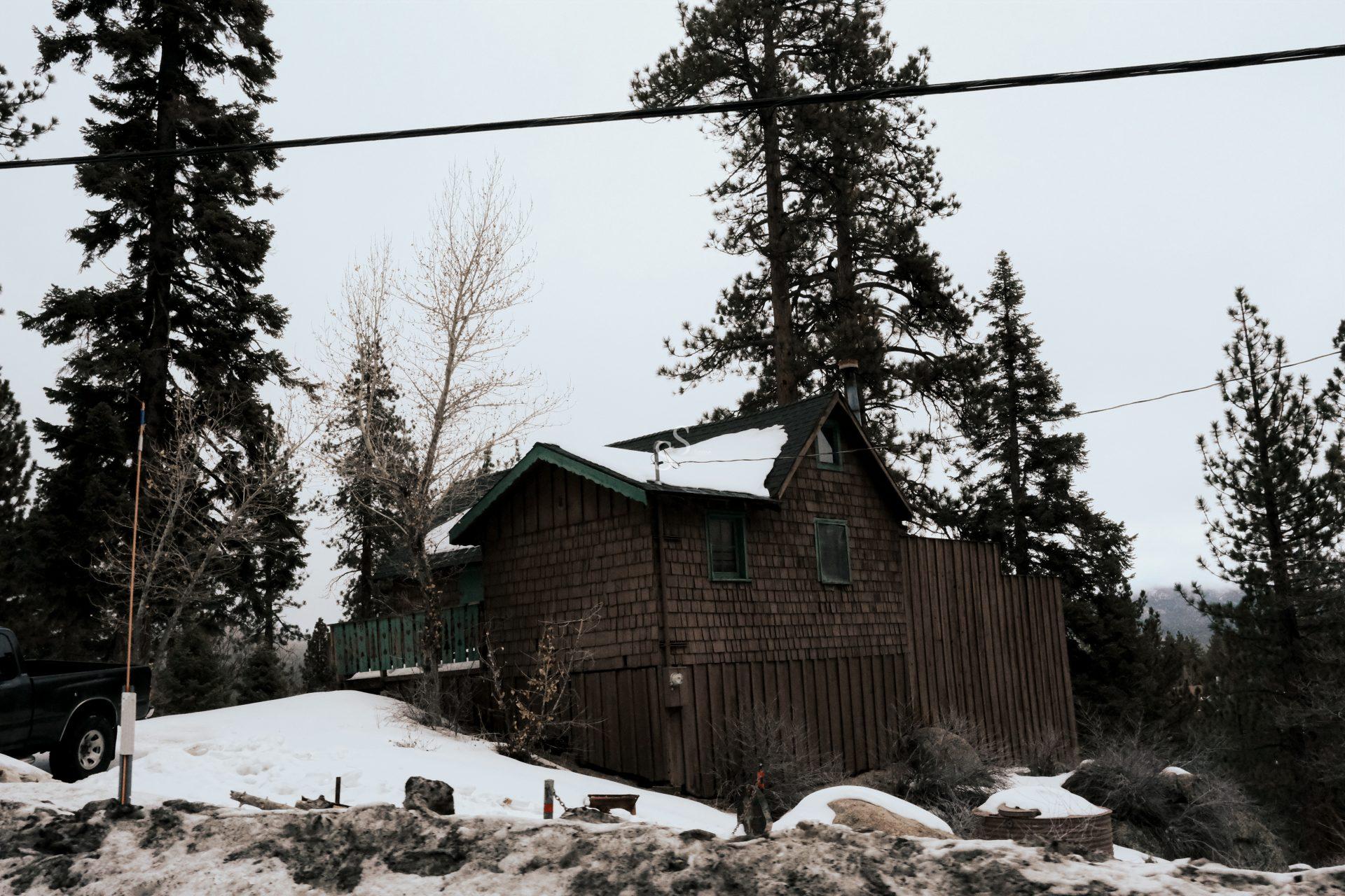 A small wooden cabin with green trim sits on a snowy hillside surrounded by tall pine trees. Power lines run overhead, and a truck is partially visible on the left. The sky is overcast.