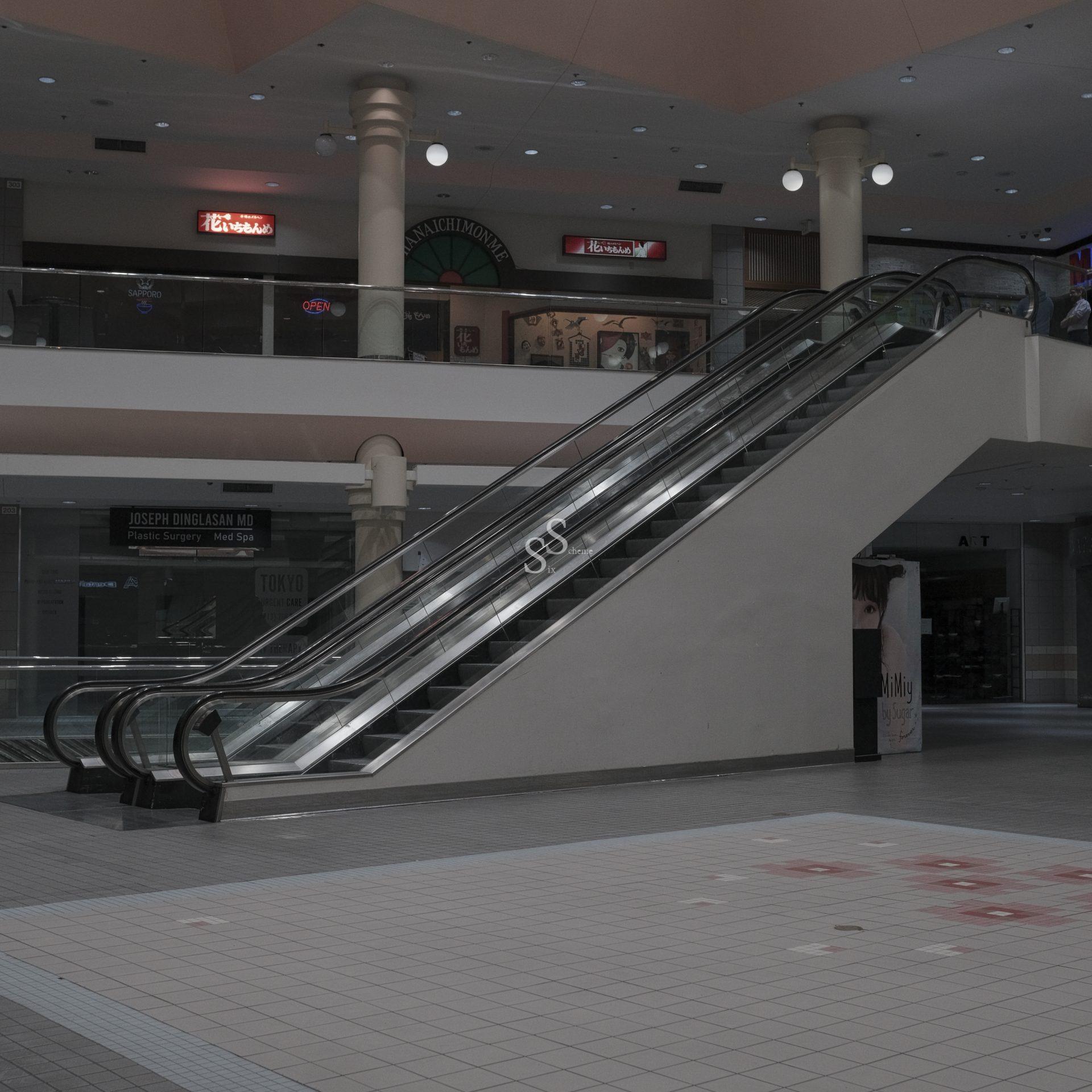 An ascending escalator cuts through the low light of an otherwise empty mall interior, passing closed stores and rising above a deserted tiled floor.