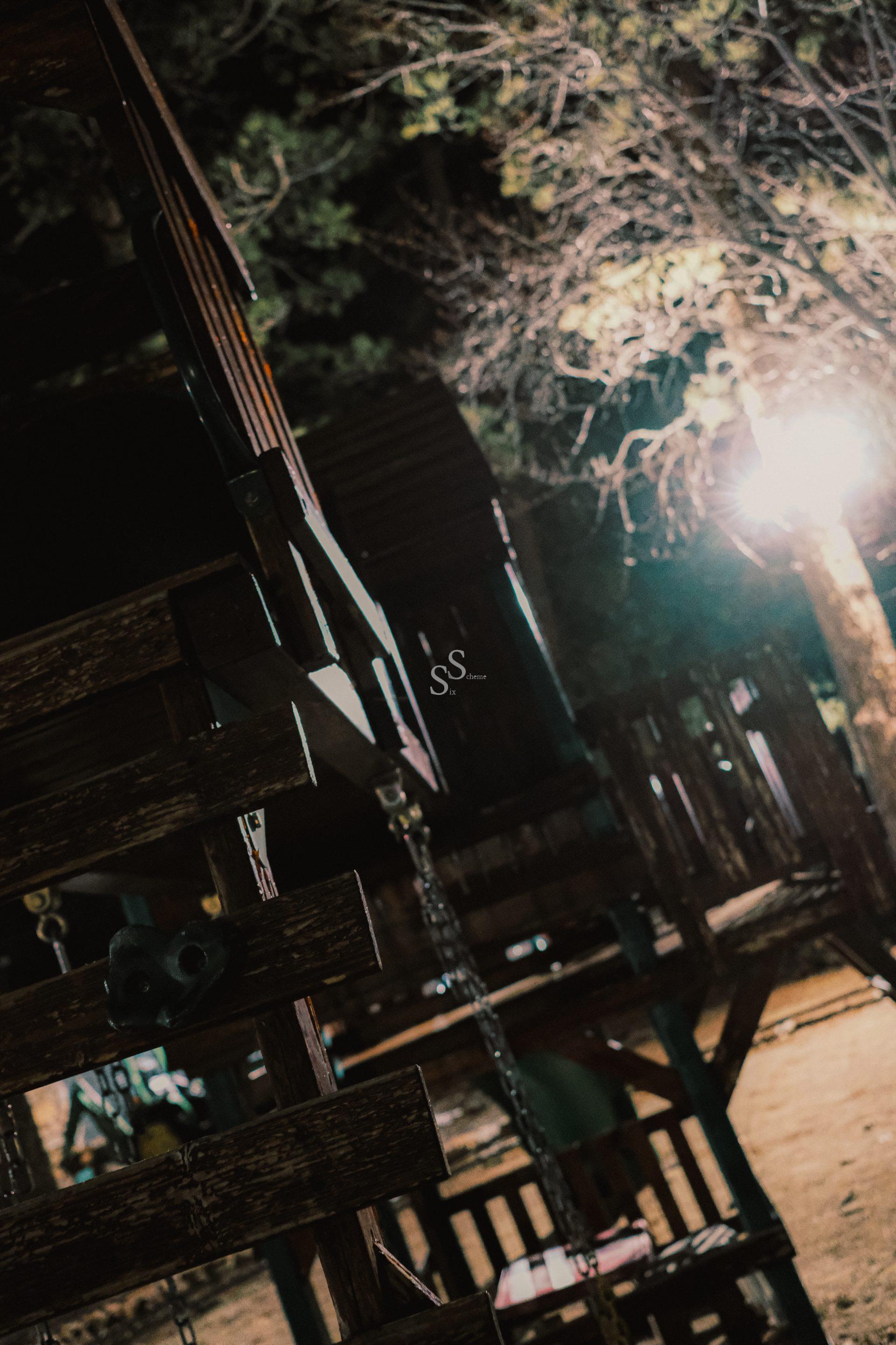 A dimly lit playground at night with wooden structures, chains, and trees in the background. A bright light source shines near the trees, creating strong shadows and an eerie atmosphere.