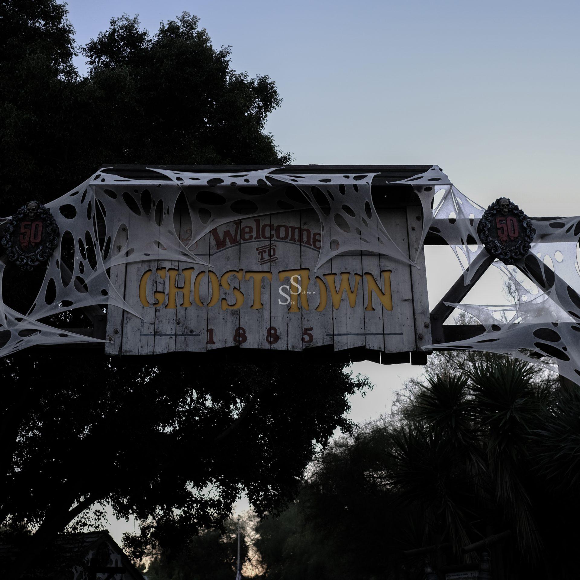 A wooden sign reading Welcome to Ghost Town 1885 is decorated with fake spider webs and two circular emblems with the number 50. Trees and a dusky sky are visible in the background.