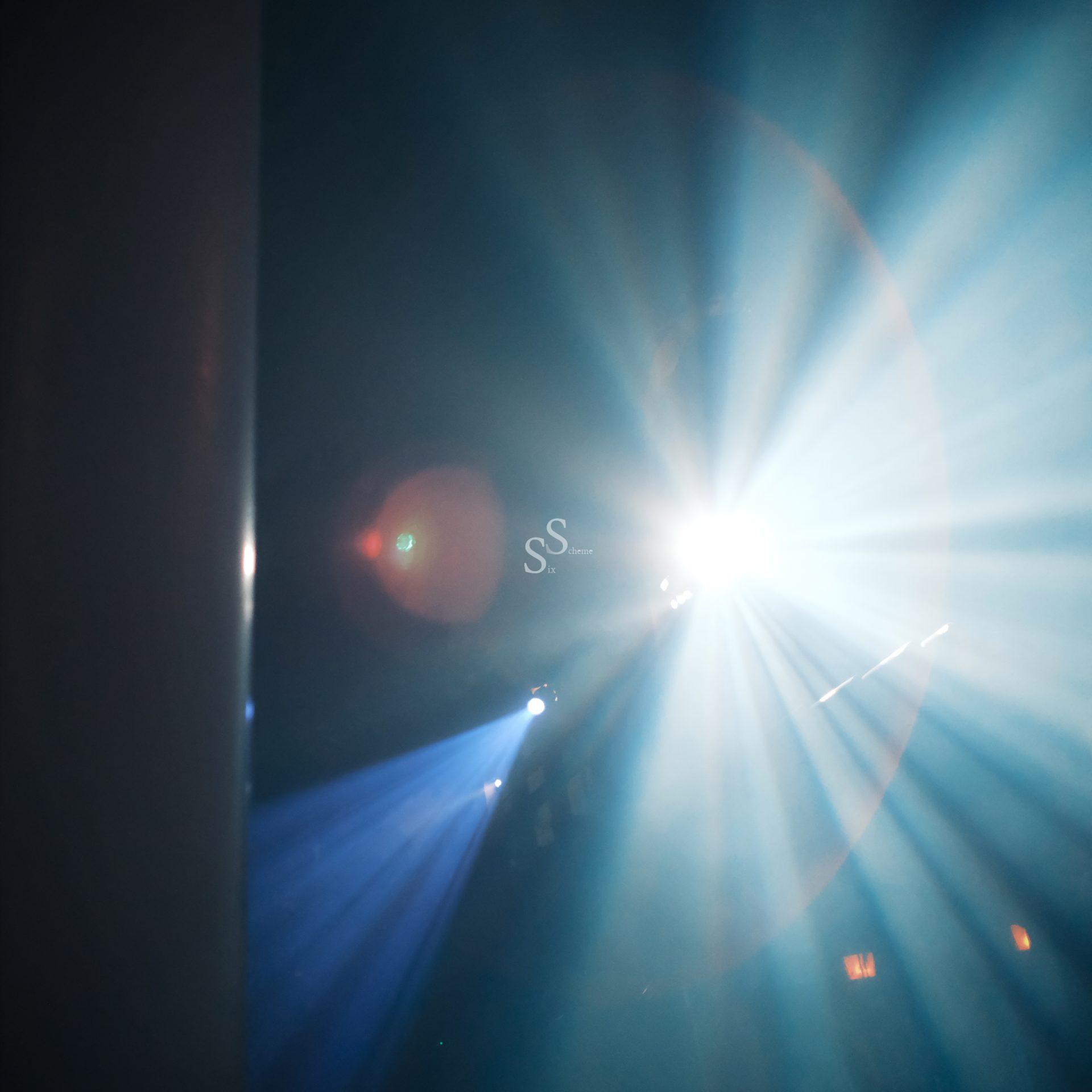 A bright white spotlight shines through haze, creating blue and white rays that fan out in the darkness. A slight lens flare and a vertical object are visible on the left side of the image.