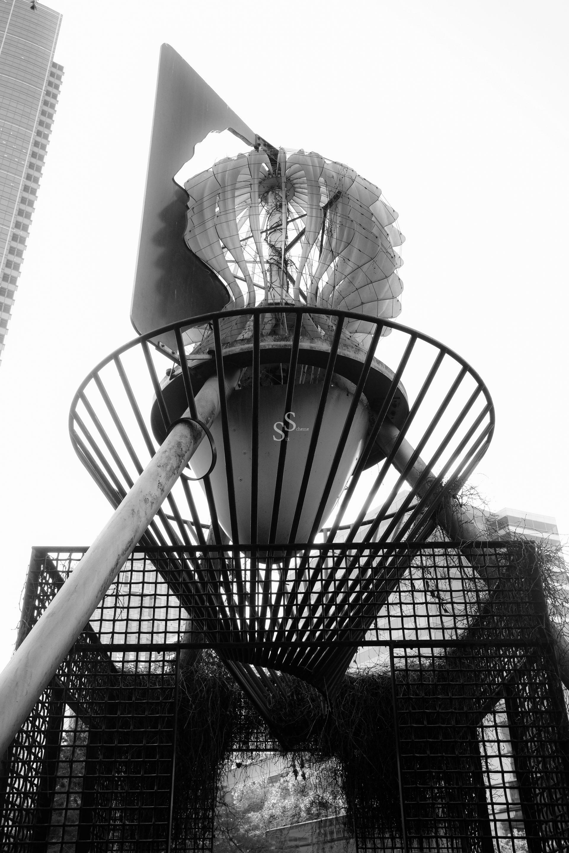 Black and white photo of a modern metal sculpture with abstract, geometric shapes, set against tall buildings. The structure has a circular base, vertical bars, and a twisted, perforated metal top. View is from a low angle.