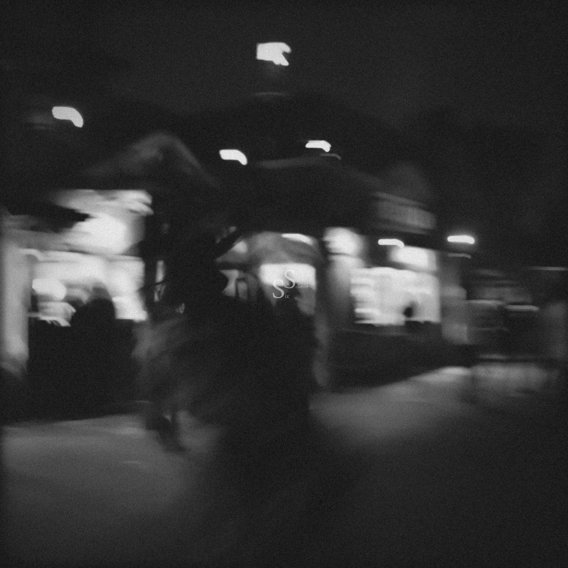 Blurry black and white photo of a night scene with glowing lights from storefronts or stalls, shadowy figures walking, and an overall hazy, indistinct atmosphere.