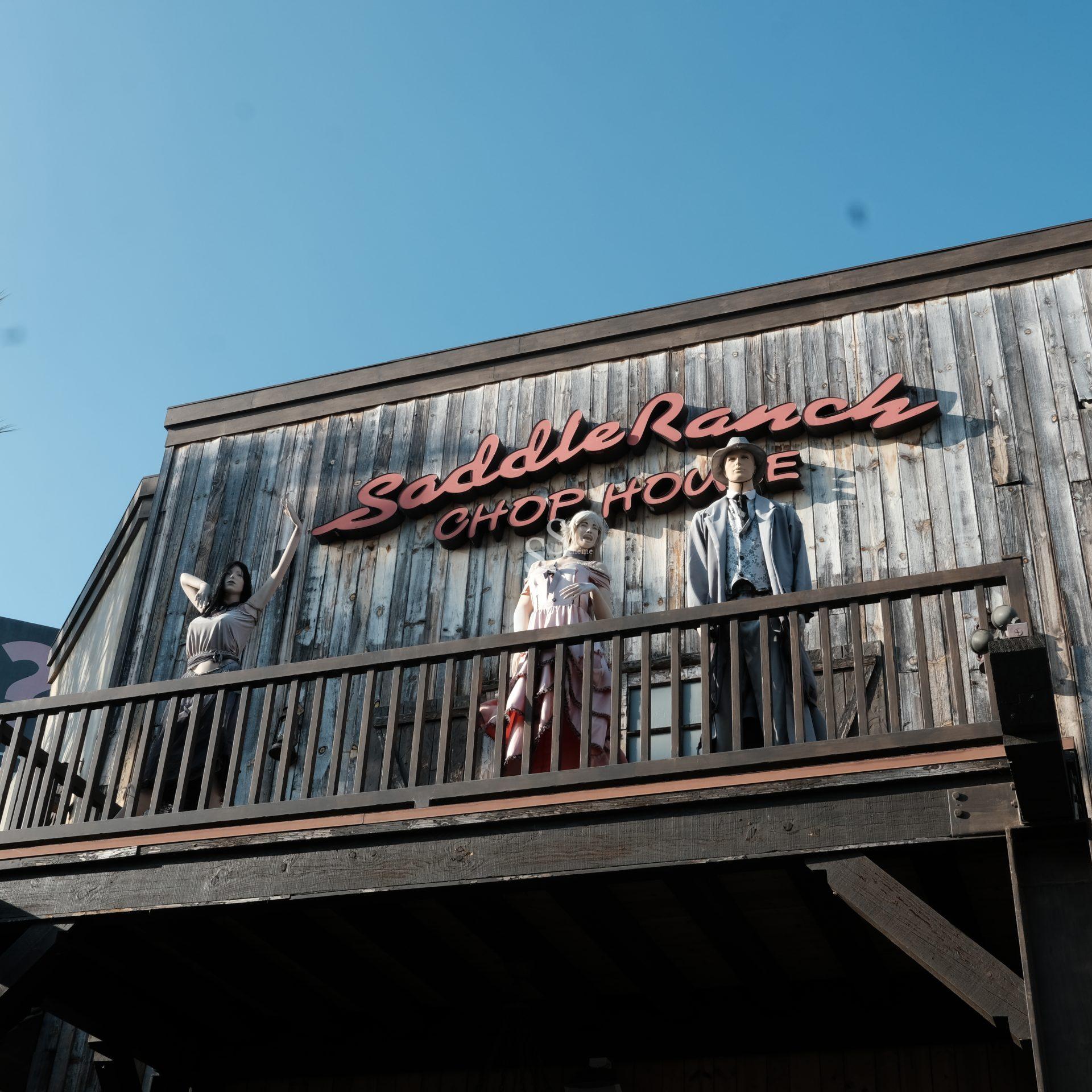 Three mannequins dressed in western attire stand on a wooden balcony under a red Saddle Ranch Chop House sign, attached to a rustic wooden building against a clear blue sky.
