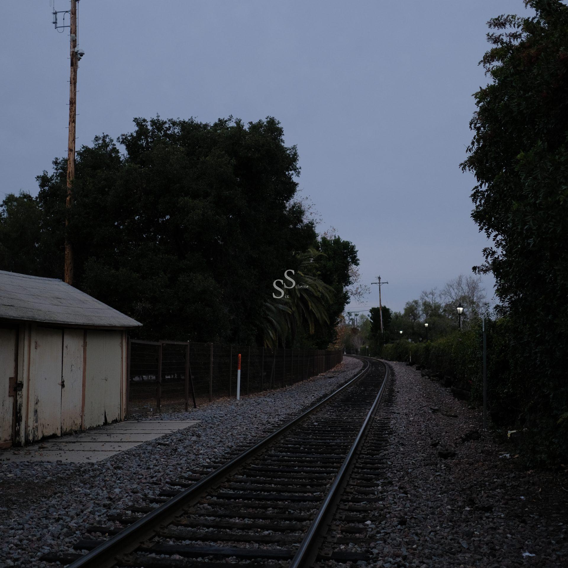 A railroad track curves to the right, bordered by trees and a fenced area with a small, weathered building on the left, under a cloudy, dimly lit sky.