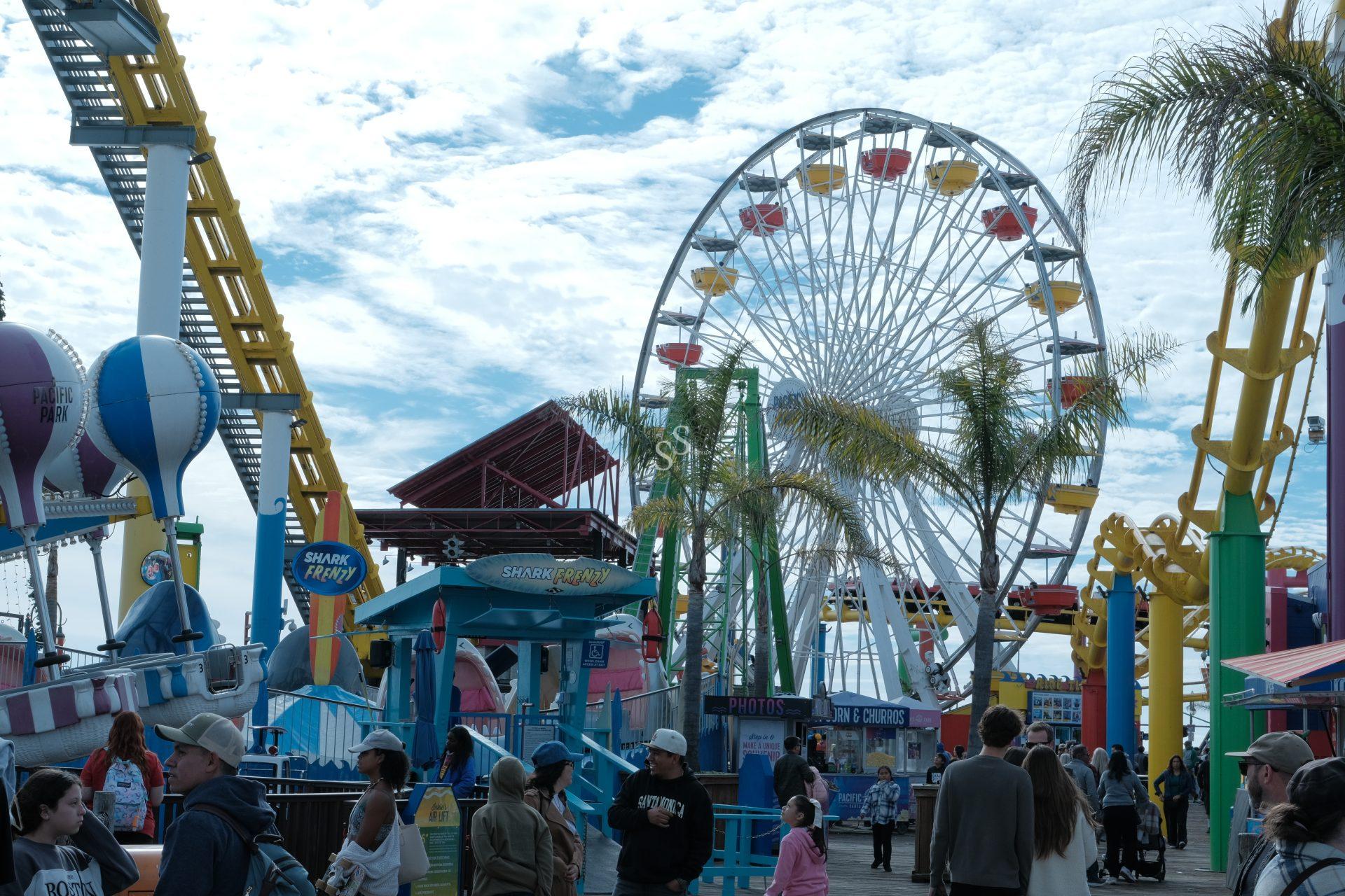 People walk along a busy amusement park boardwalk with a large Ferris wheel, roller coaster tracks, palm trees, and colorful attractions under a partly cloudy sky.