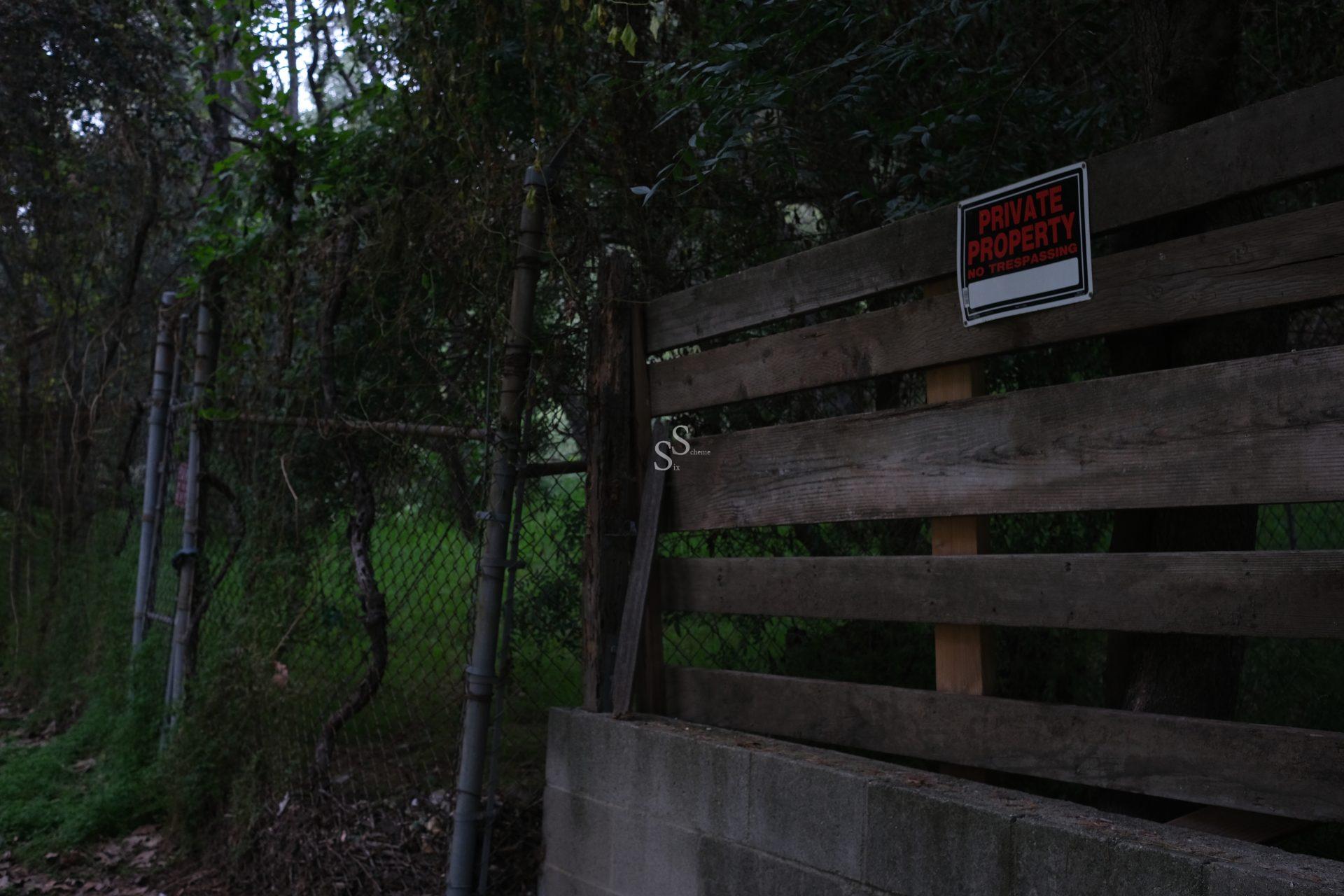 A wooden fence with a Private Property sign is in front of a chain-link fence and dense trees. The area behind the fence appears green and overgrown. The lighting is dim, suggesting early evening or shade.