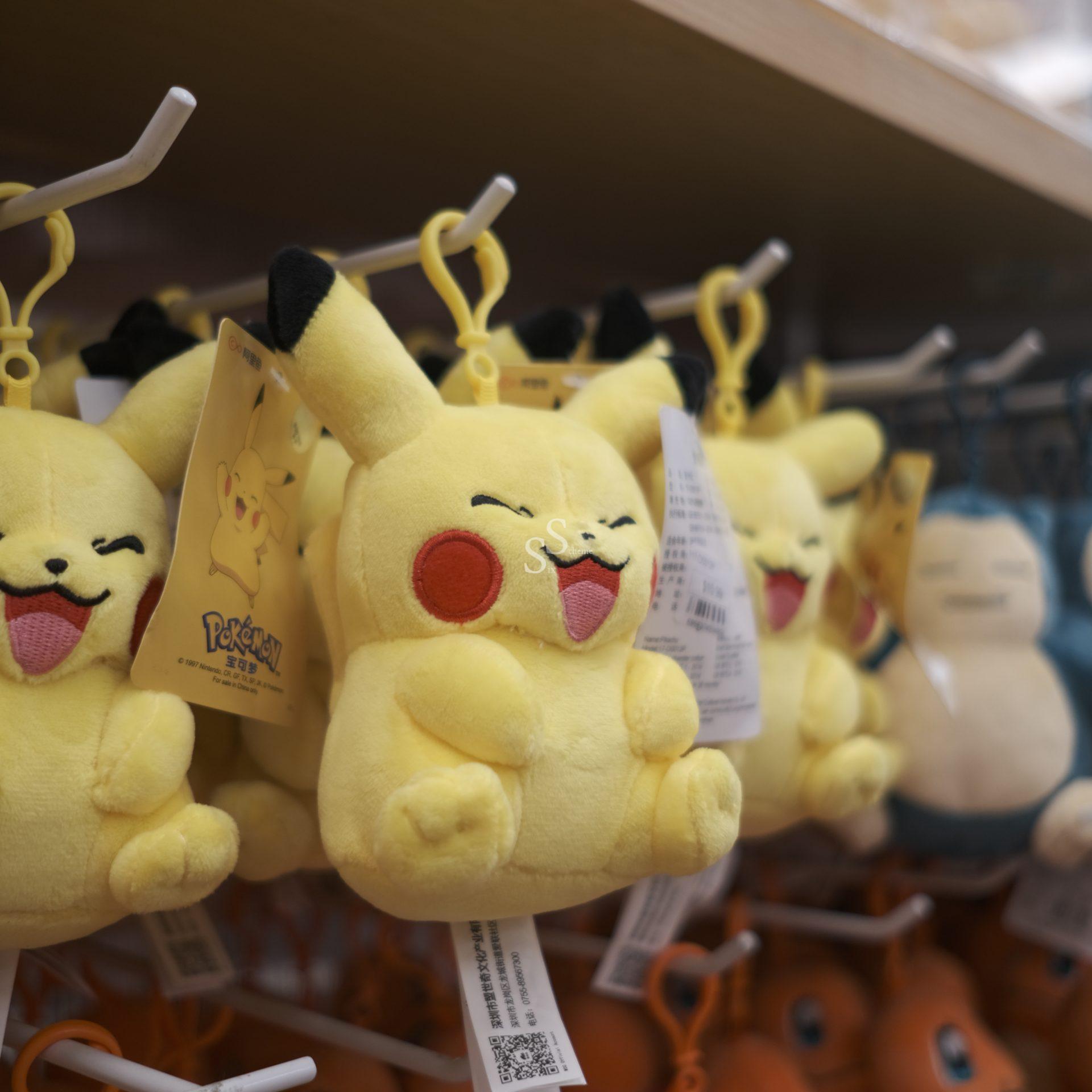 Plush Pikachu keychains with smiling faces hang on a store display rack, with more plush toys visible in the background.