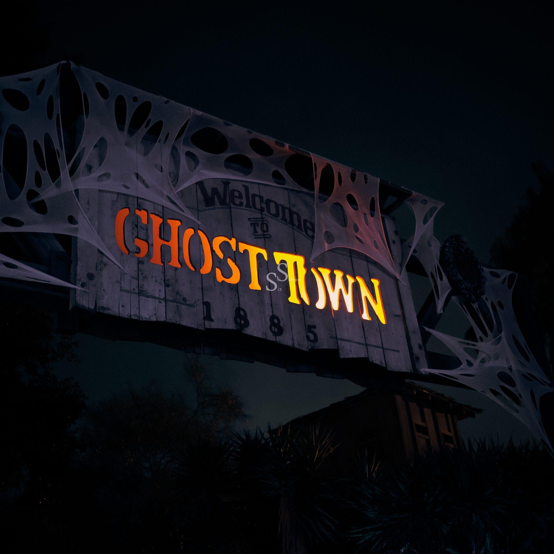 A spooky sign at night reads “Welcome to Ghost Town 1885,” lit with orange and yellow letters and draped in fake cobwebs. The background is dark with faint outlines of trees and foliage.