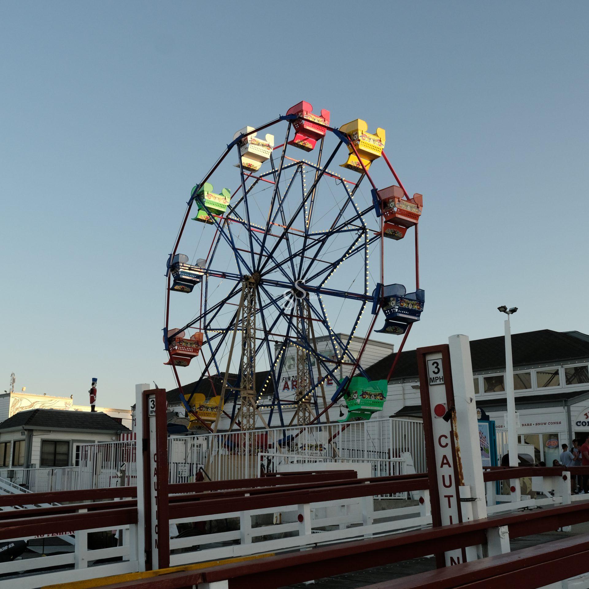 A colorful Ferris wheel stands against a clear blue sky at an amusement park, with red, yellow, blue, and green passenger cars. White fences and safety signs are visible in the foreground.