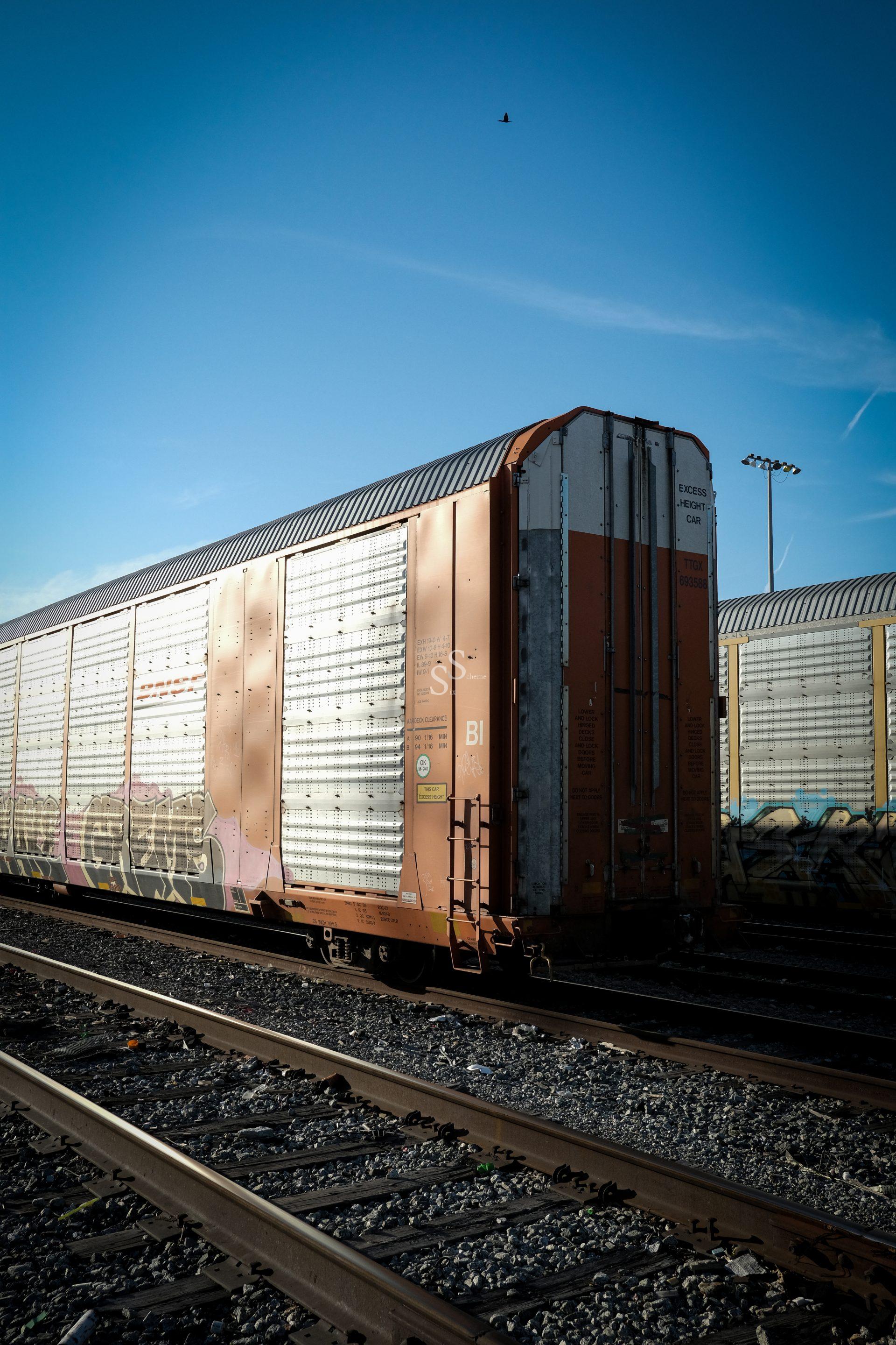 A brown and silver freight train car sits stationary on railway tracks under a clear blue sky, with sunlight casting shadows and another train car visible in the background.