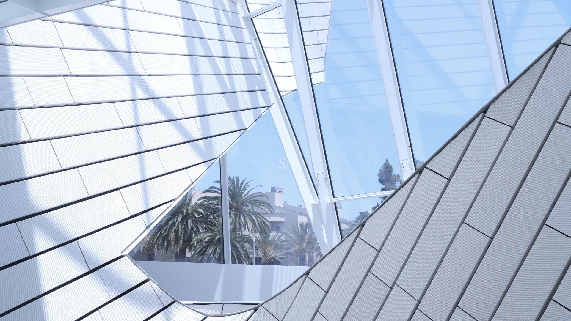Modern building interior with angled metal panels and large glass windows. Palm trees and a clear blue sky are visible outside, contrasting with the geometric architecture and soft natural light streaming in.