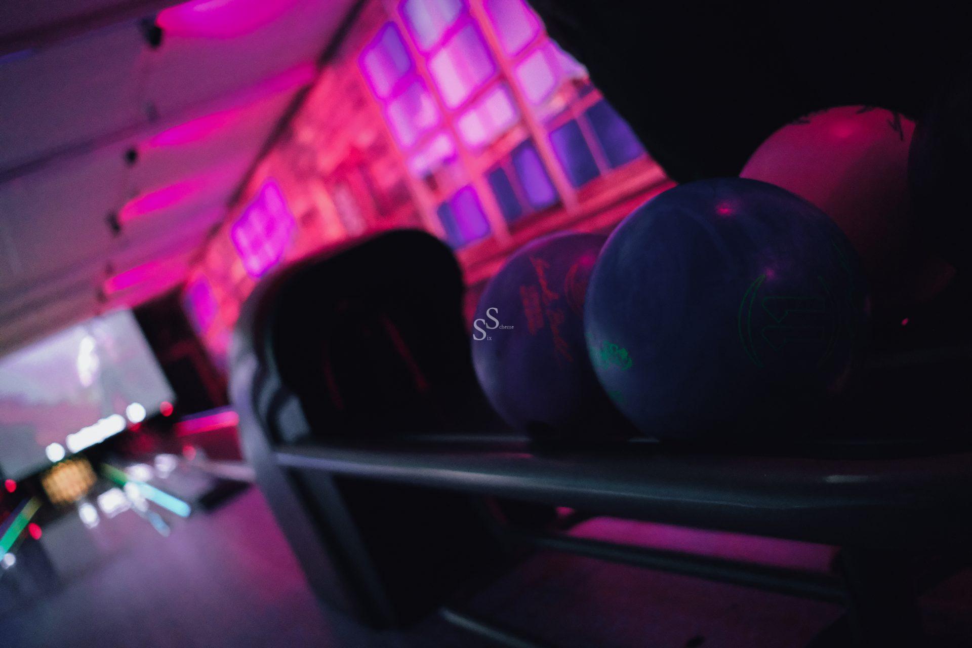 Bowling balls rest on a rack in a dimly lit bowling alley, illuminated by pink and purple neon lights, with lanes and pins visible in the background.