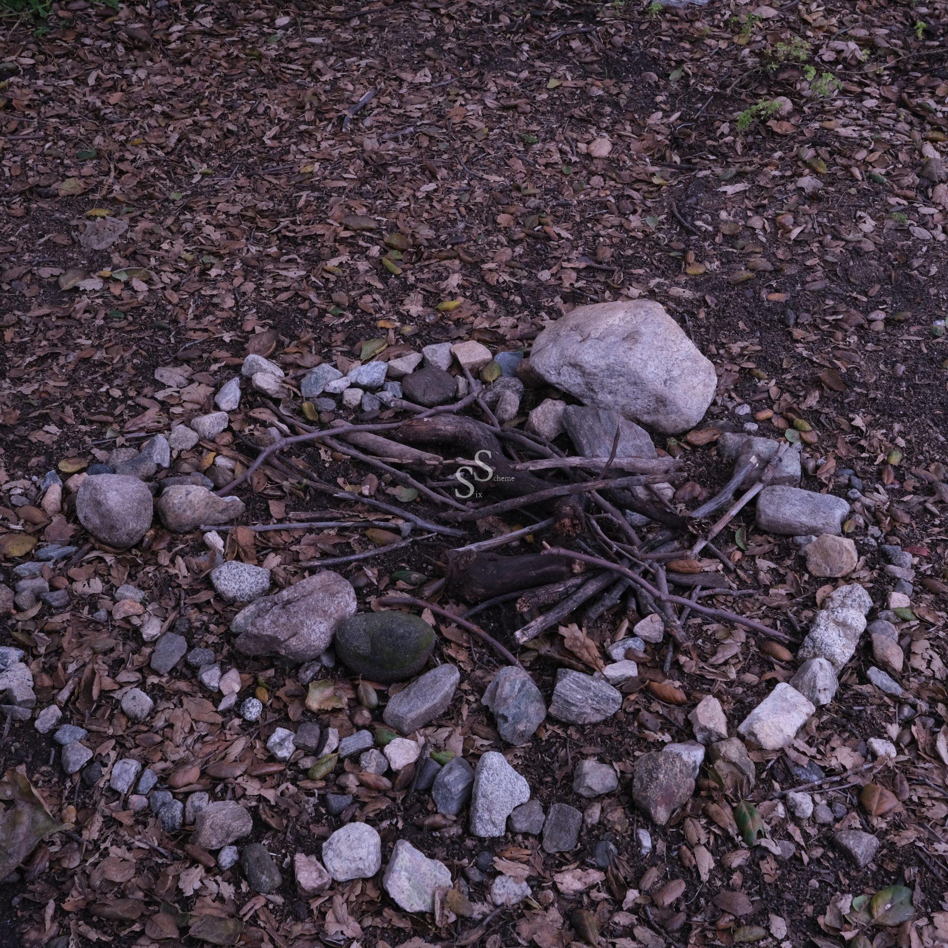 A small campfire ring made of rocks on the ground, filled with dry sticks and twigs, surrounded by fallen leaves and dirt in a wooded outdoor area.