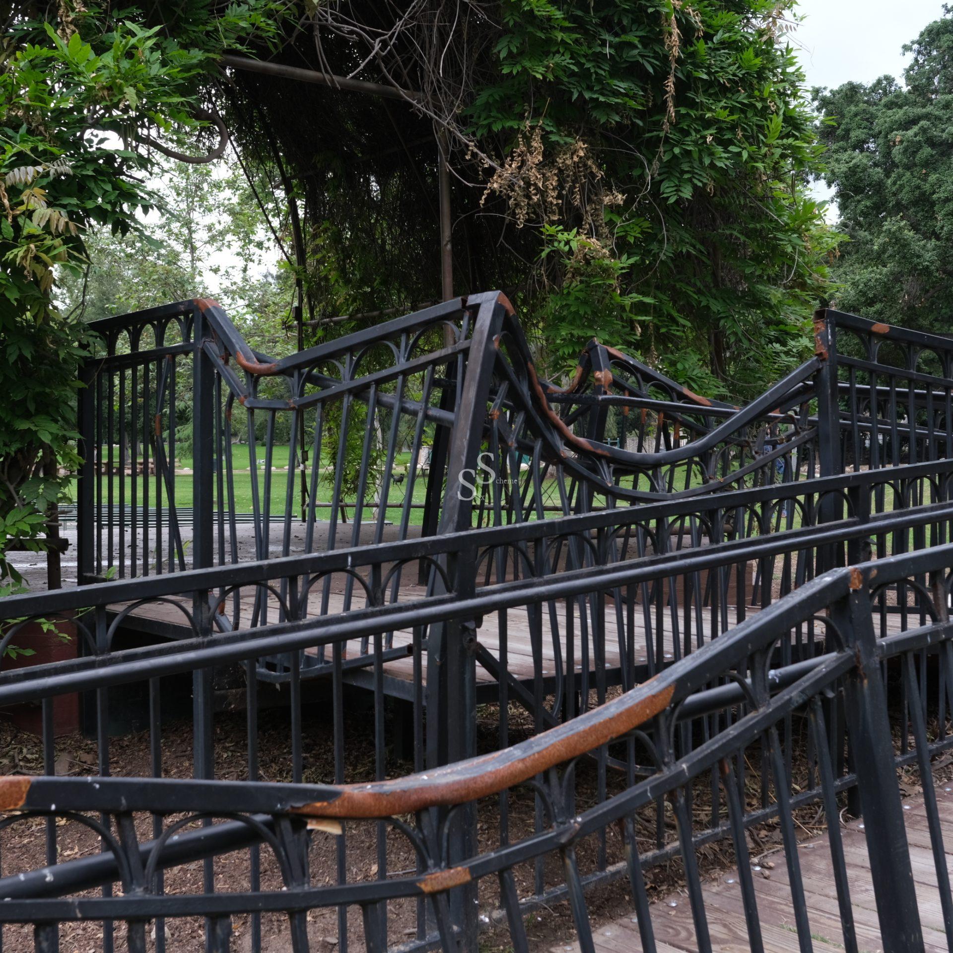 A winding wooden bridge with unusually twisted black metal railings, surrounded by green trees and climbing vines.