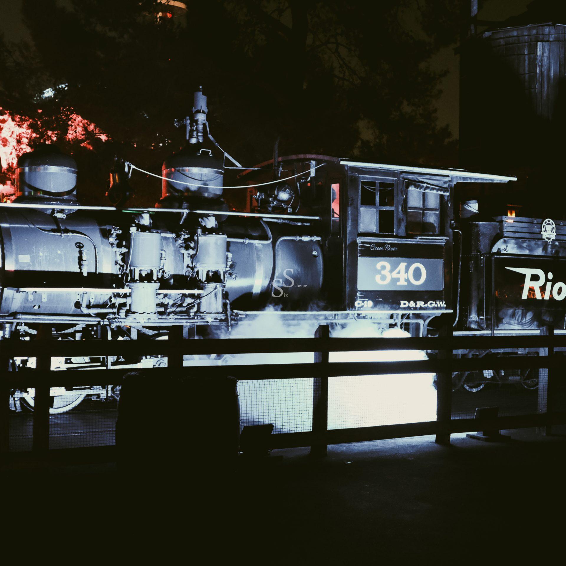 A vintage steam locomotive numbered 340 emits steam at night, illuminated by dramatic lighting. The train is behind a fence, with trees and faint red lights in the background.