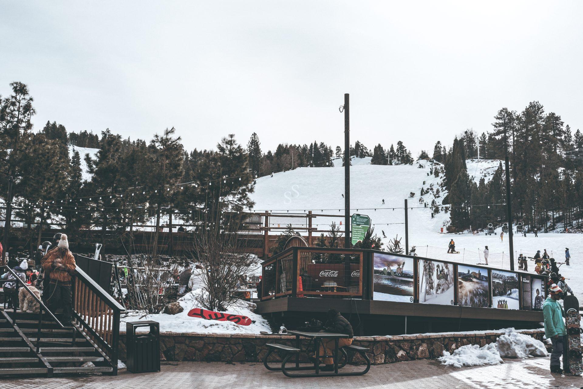 A snowy ski resort with skiers on a slope, trees in the background, people walking and standing, and a wooden deck area with tables and signs in the foreground.