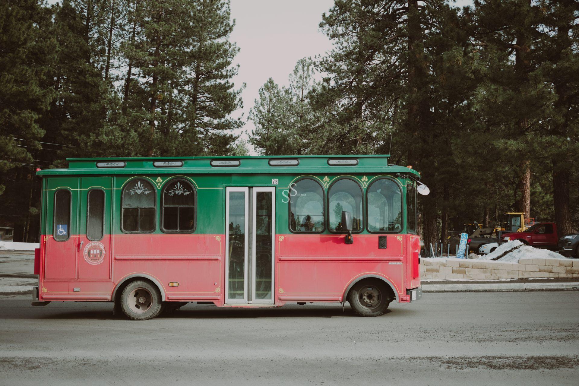 A red and green trolley bus is parked on a road, surrounded by tall pine trees and patches of snow on the ground.
