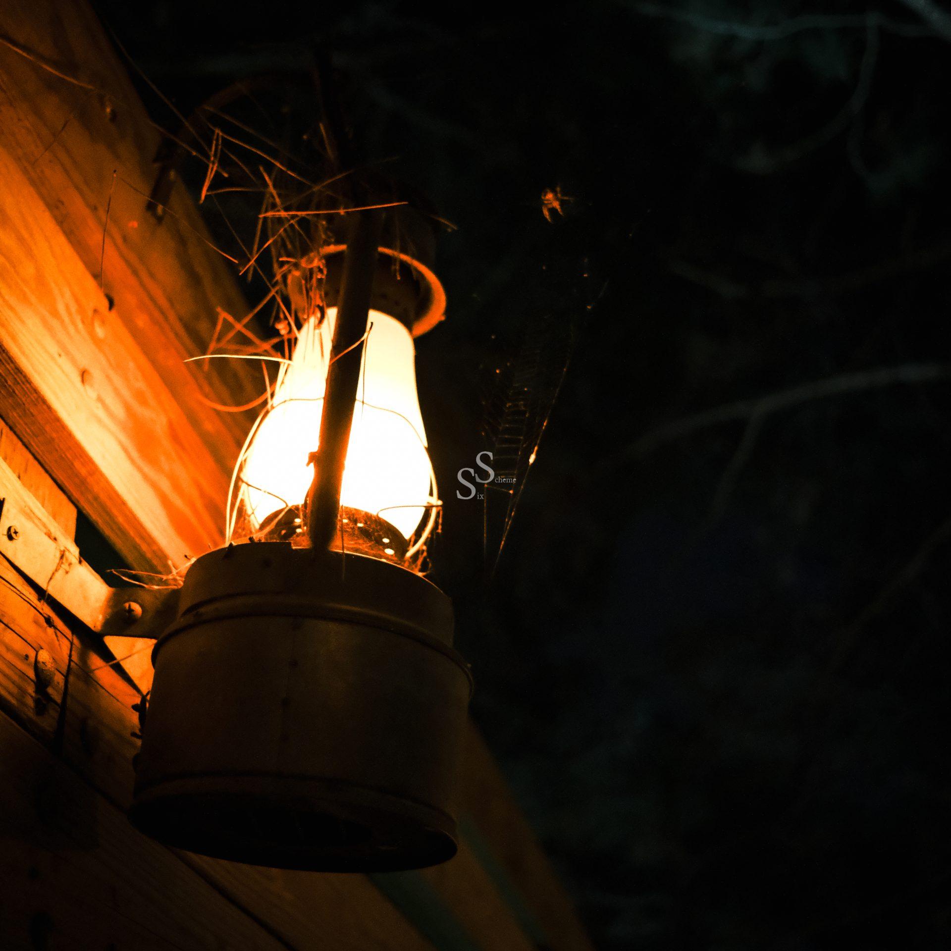 A lit lantern mounted on a wooden wall glows warmly at night, illuminating cobwebs and a spider above it, with a dark, shadowy background.