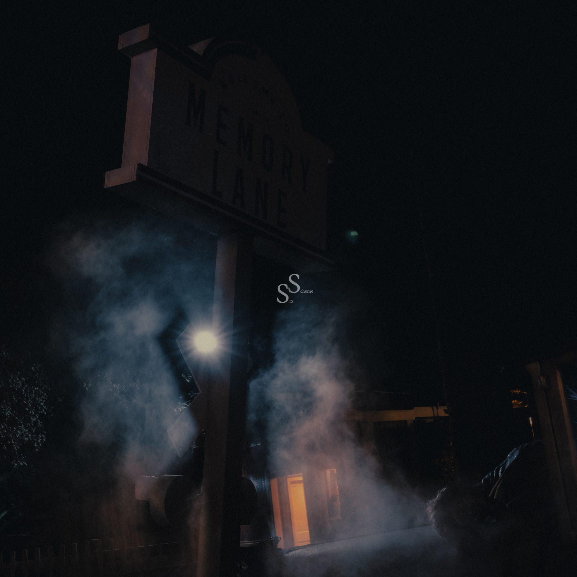 A dimly lit street sign reading “Memory Lane” stands in the dark, surrounded by mist and illuminated by a bright light, with a building faintly visible in the background.