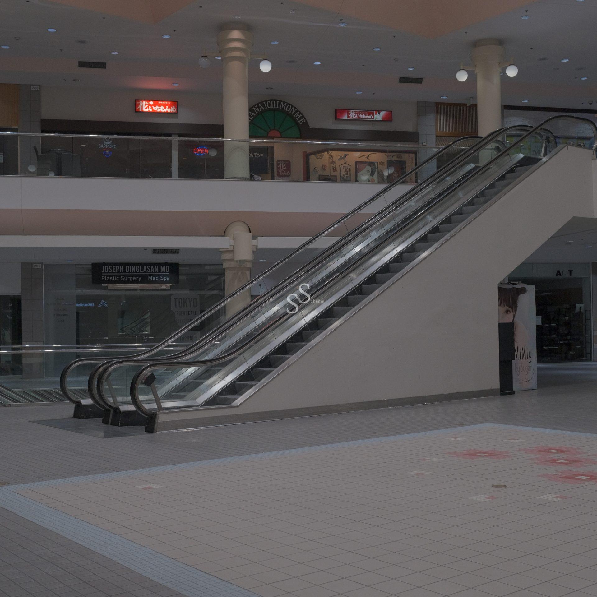 An empty mall interior with an escalator going up, dim lighting, closed stores, and deserted tiled flooring.