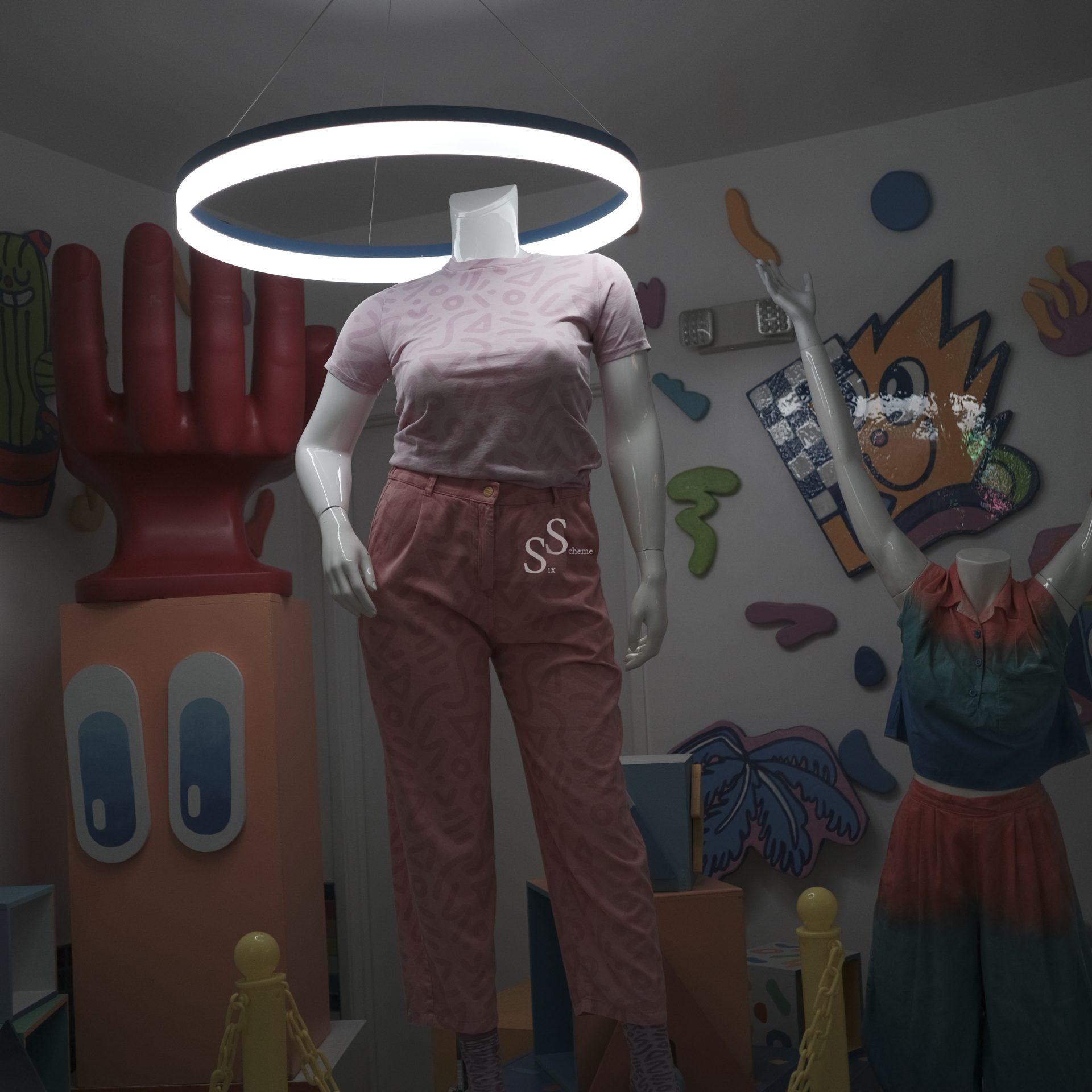 A headless mannequin wearing a pink patterned shirt and pants stands under a circular light, surrounded by colorful abstract art, a red hand sculpture, and another dressed mannequin in a vibrant room.
