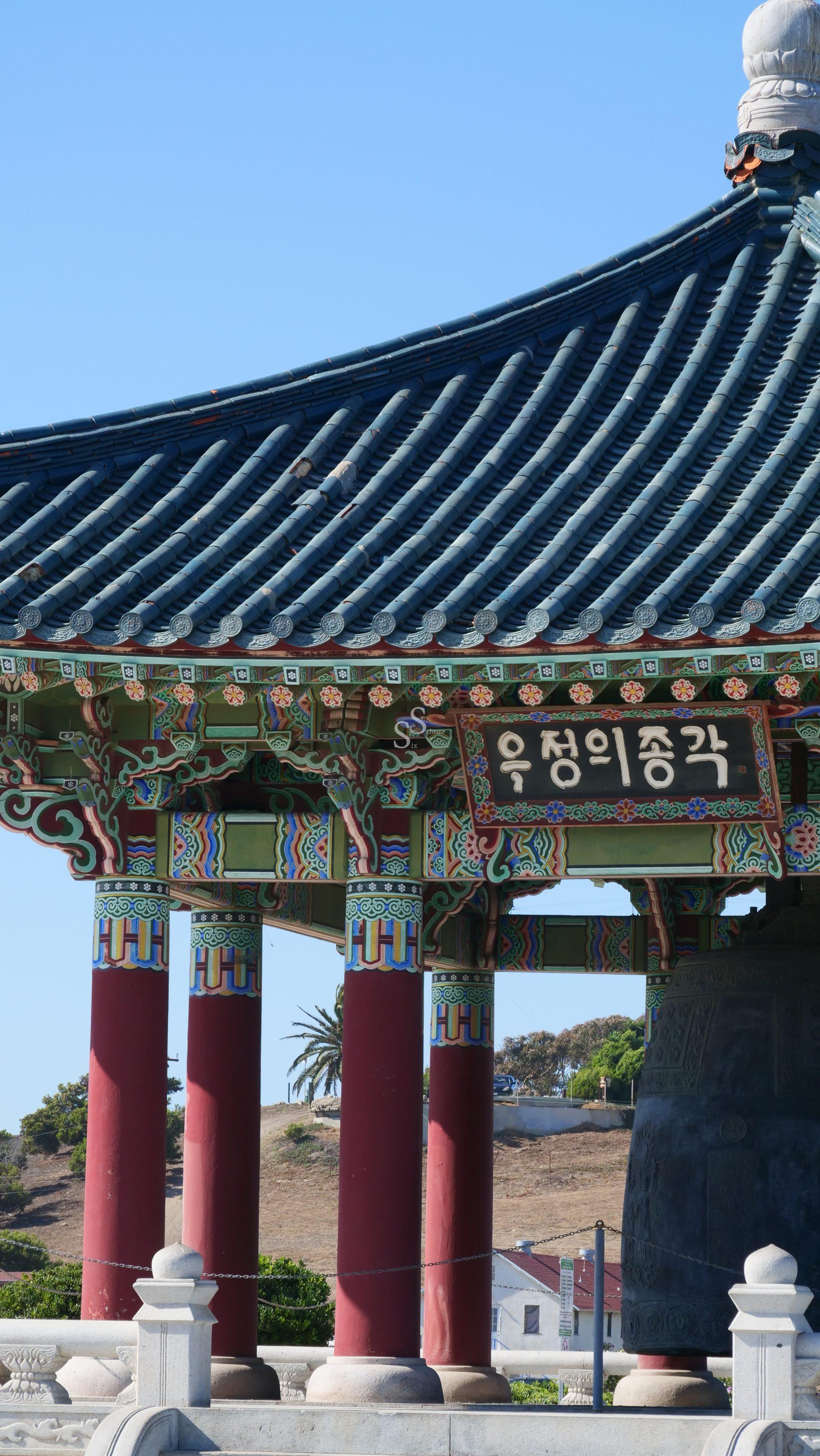 A close-up of a traditional Korean pagoda with ornate painted patterns, red columns, and a sign with Korean writing. Palm trees and clear blue sky are visible in the background.