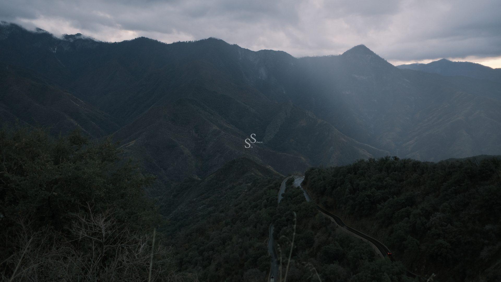 A winding road curves through forested green mountains under a cloudy, overcast sky with sunlight breaking through, highlighting the misty peaks in the distance.