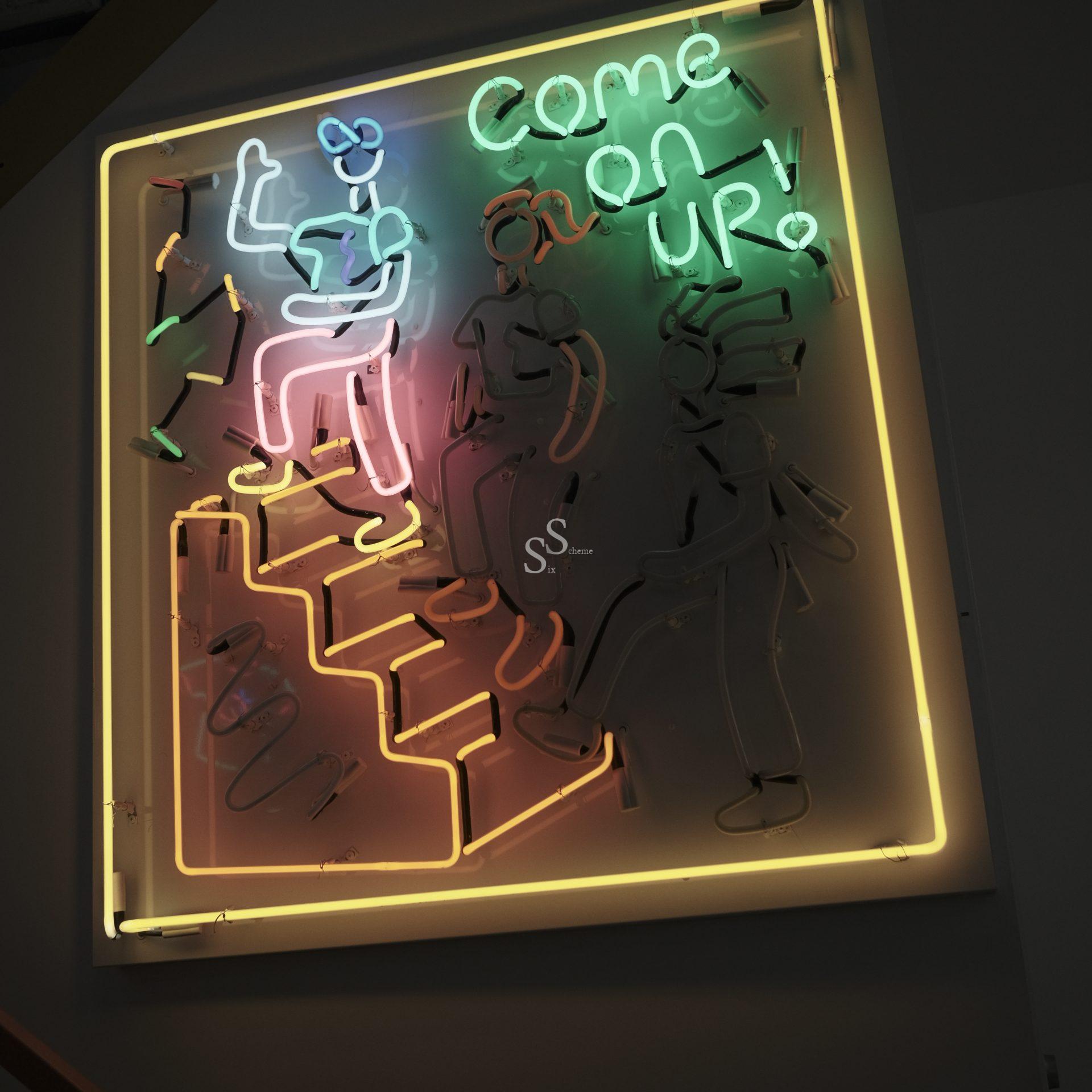 Neon sign showing three outlined figures walking up stairs, with the words “COME ON UP!” in green neon above them. The artwork is outlined with a yellow neon border against a dark background.