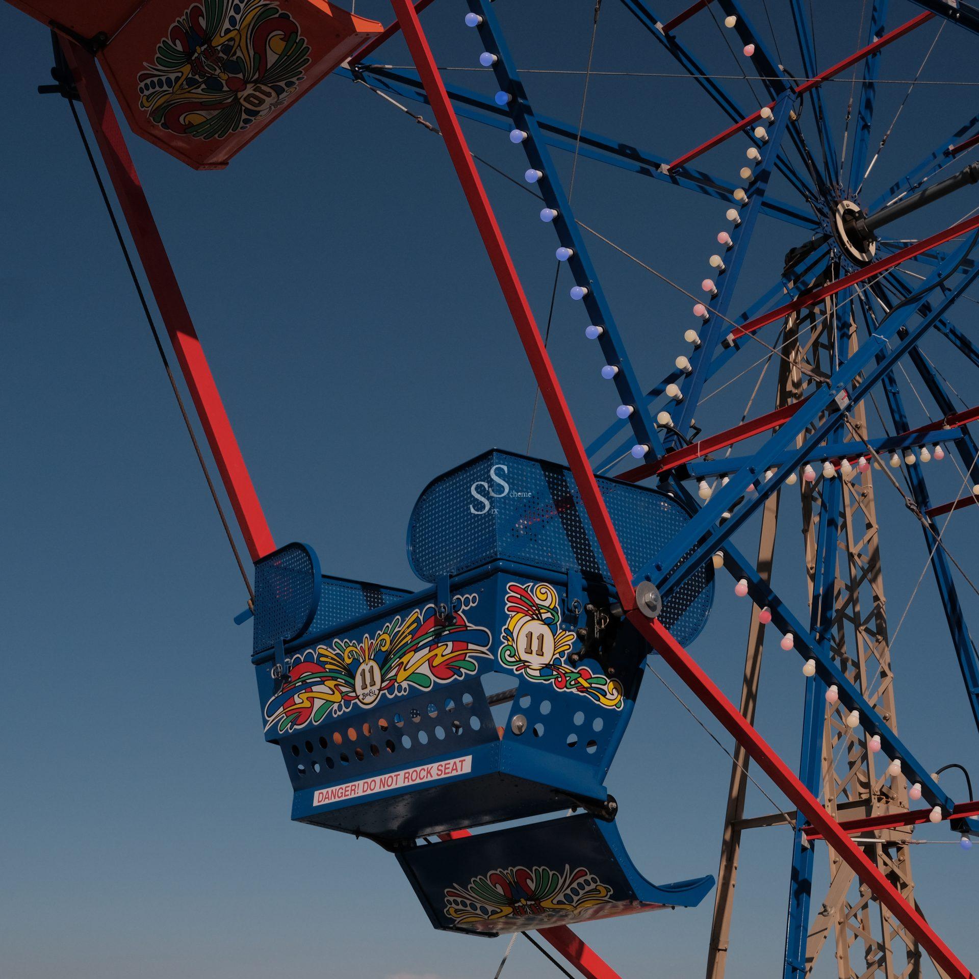 A close-up of a blue Ferris wheel gondola decorated with colorful patterns and the number 11, set against a clear blue sky. A warning label reads, “DANGER! DO NOT ROCK SEAT.”.