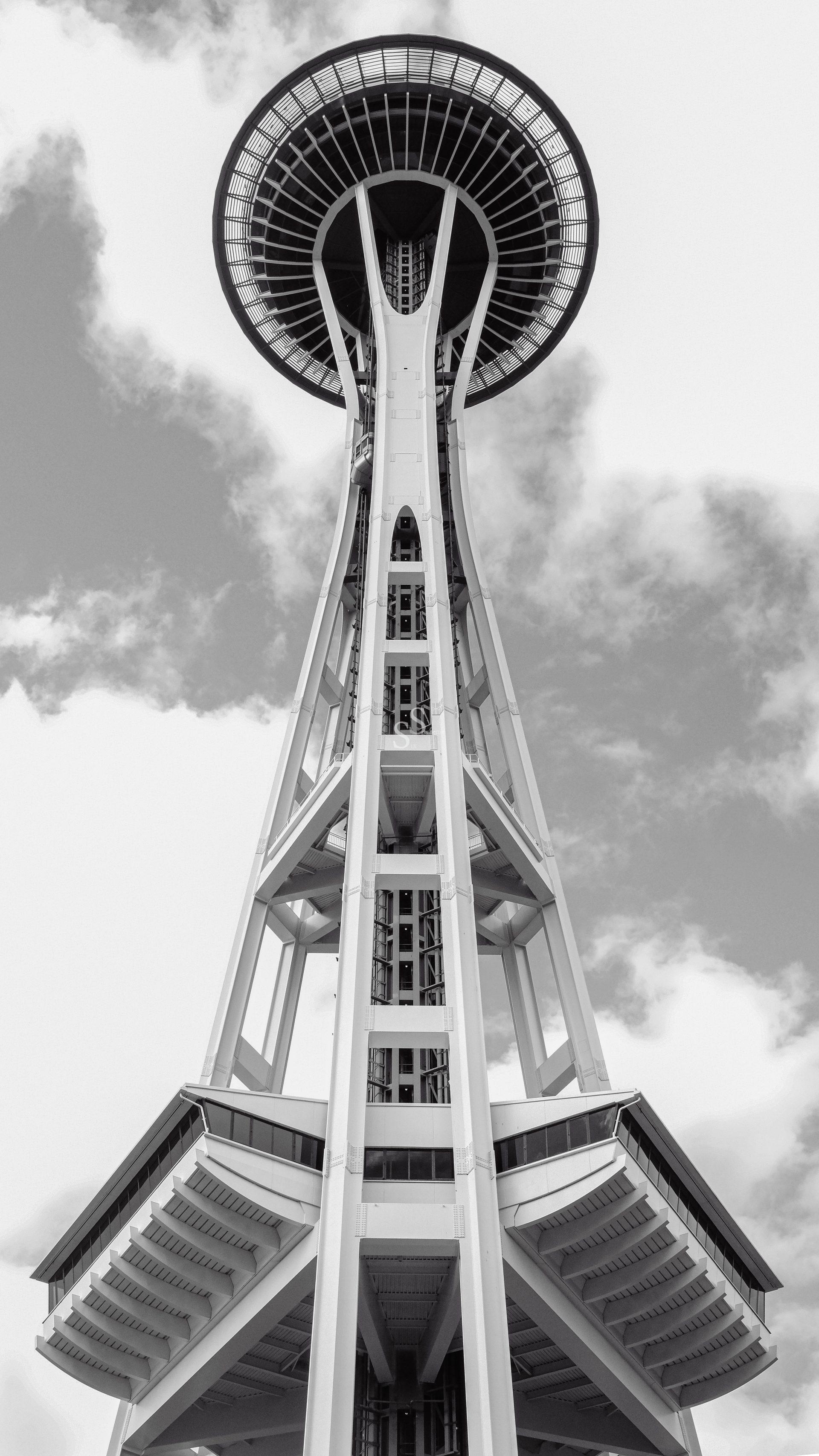Black and white photo of the Space Needle in Seattle, viewed from below, with clouds in the sky behind the iconic tower’s structure.