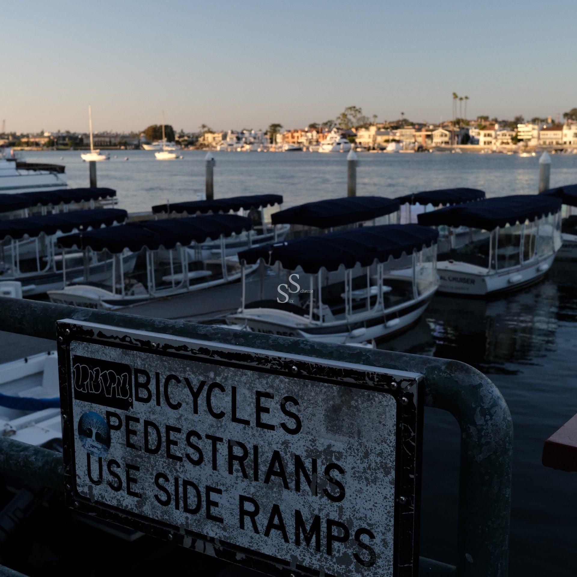 A weathered sign by a marina reads Bicycles Pedestrians Use Side Ramps with a sticker of a tree symbol. Covered boats are docked in the water, and houses are visible across the bay under a clear sky at sunset.