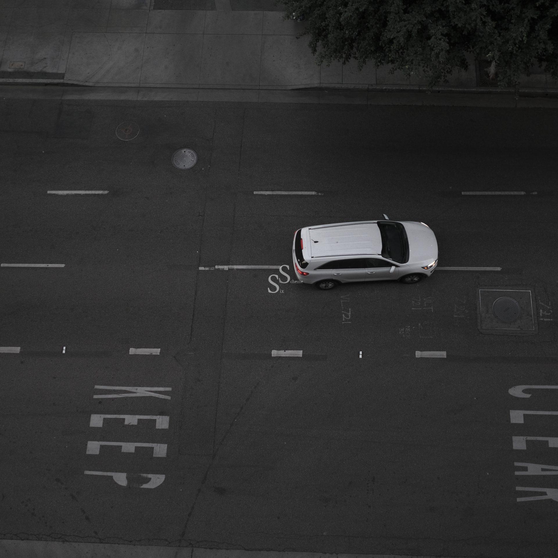 A white car drives down an empty street with the word KEEP CLEAR painted on the asphalt. The scene is mostly gray, with trees visible in the upper right corner.