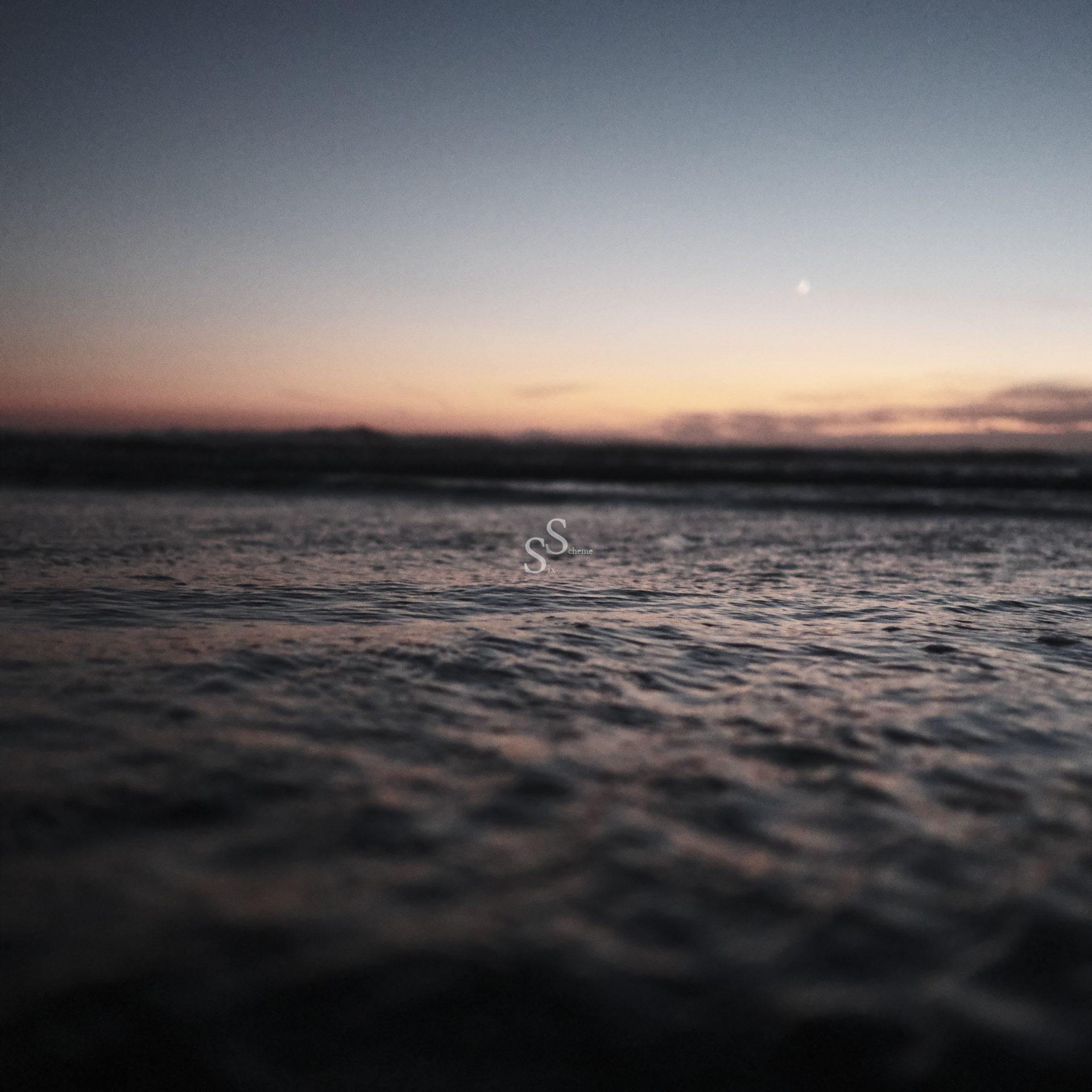 A calm ocean at dusk with gentle waves in the foreground and a soft, pastel-colored sky transitioning from orange to blue as the sun sets. A crescent moon is visible in the distance above the horizon.