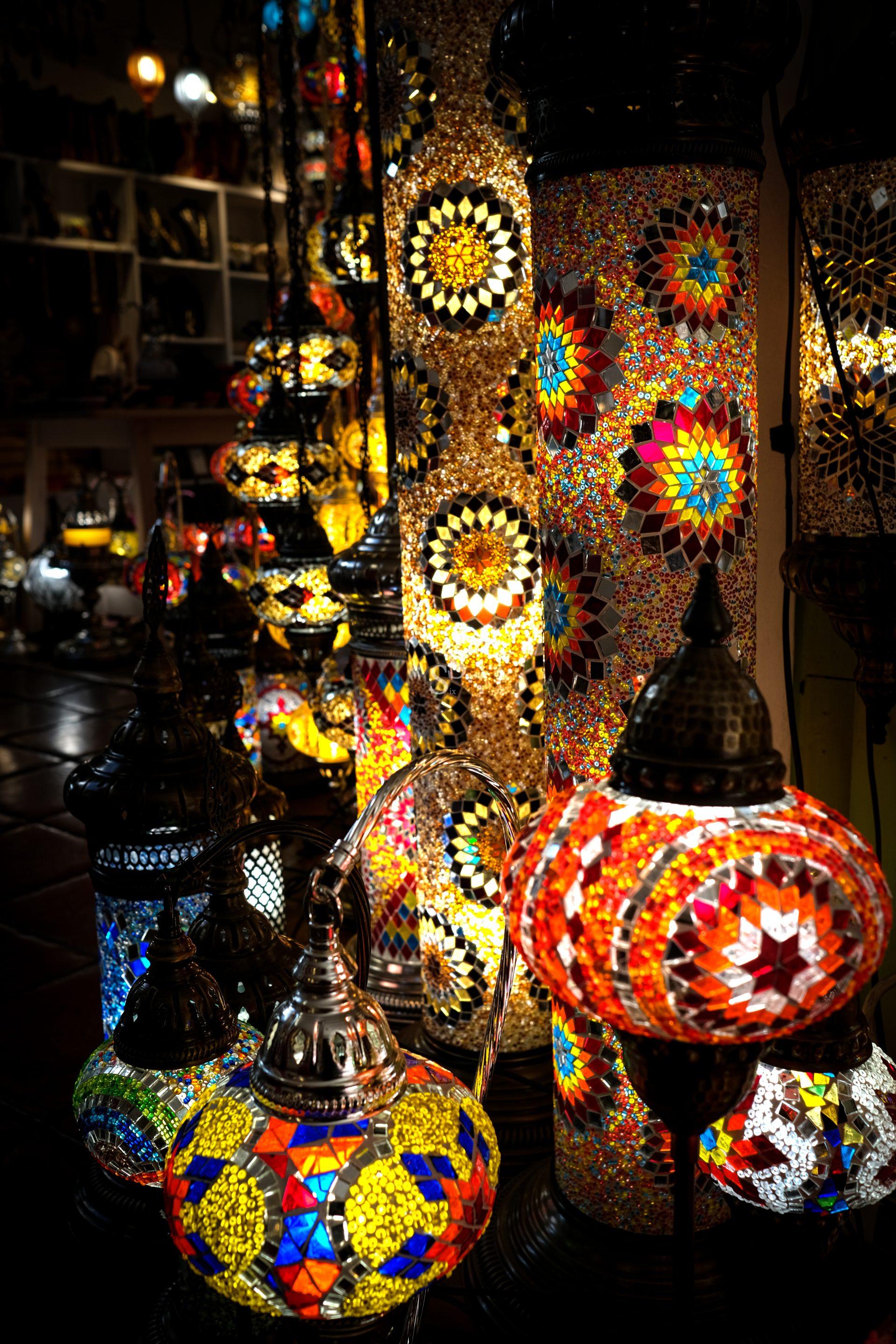 Colorful mosaic glass Turkish lamps and lanterns are displayed closely together, glowing warmly in various patterns and colors against a dimly lit background.