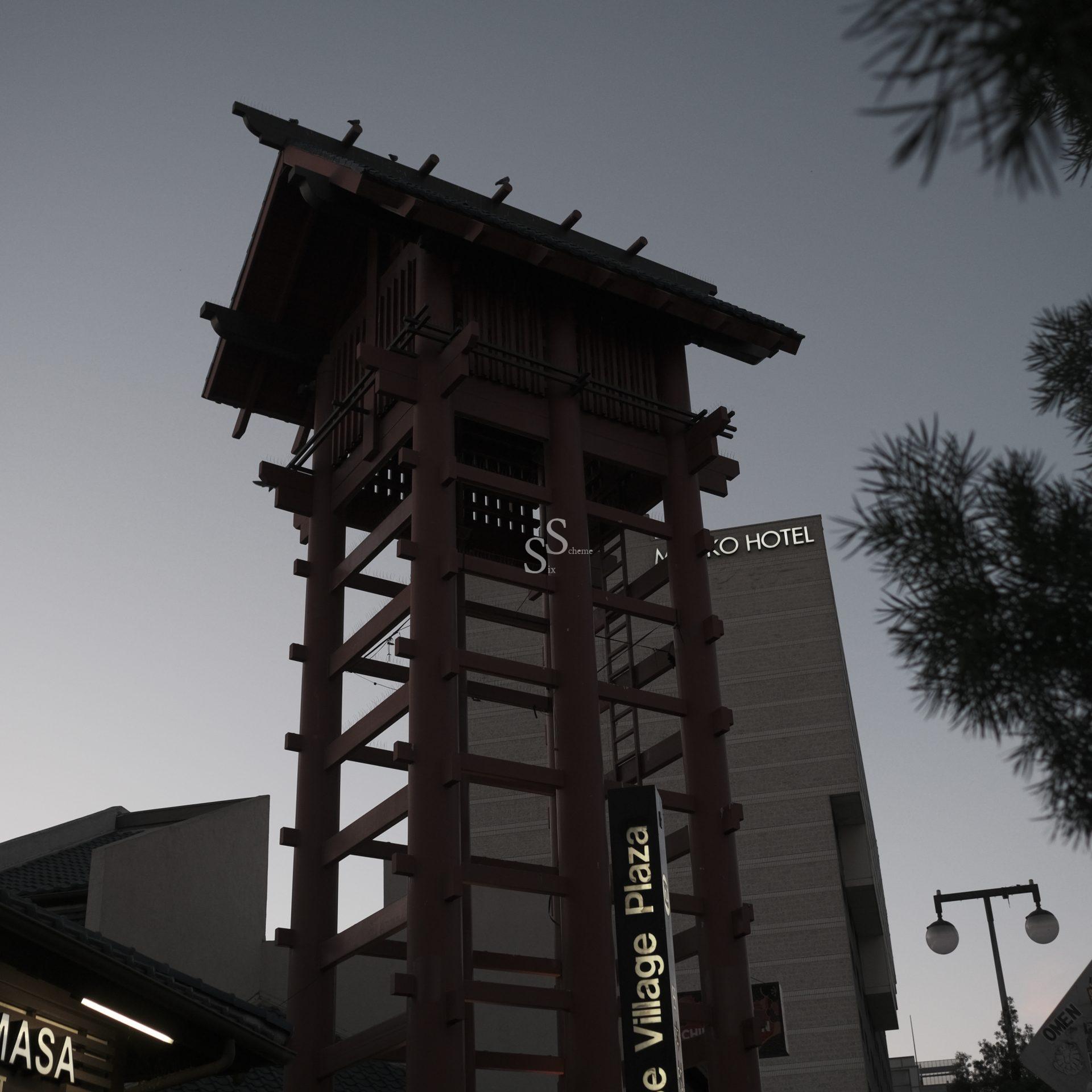 A tall wooden tower with a traditional Japanese architectural style stands in front of modern buildings at dusk, with signs reading Village Plaza and HOTEL visible nearby.