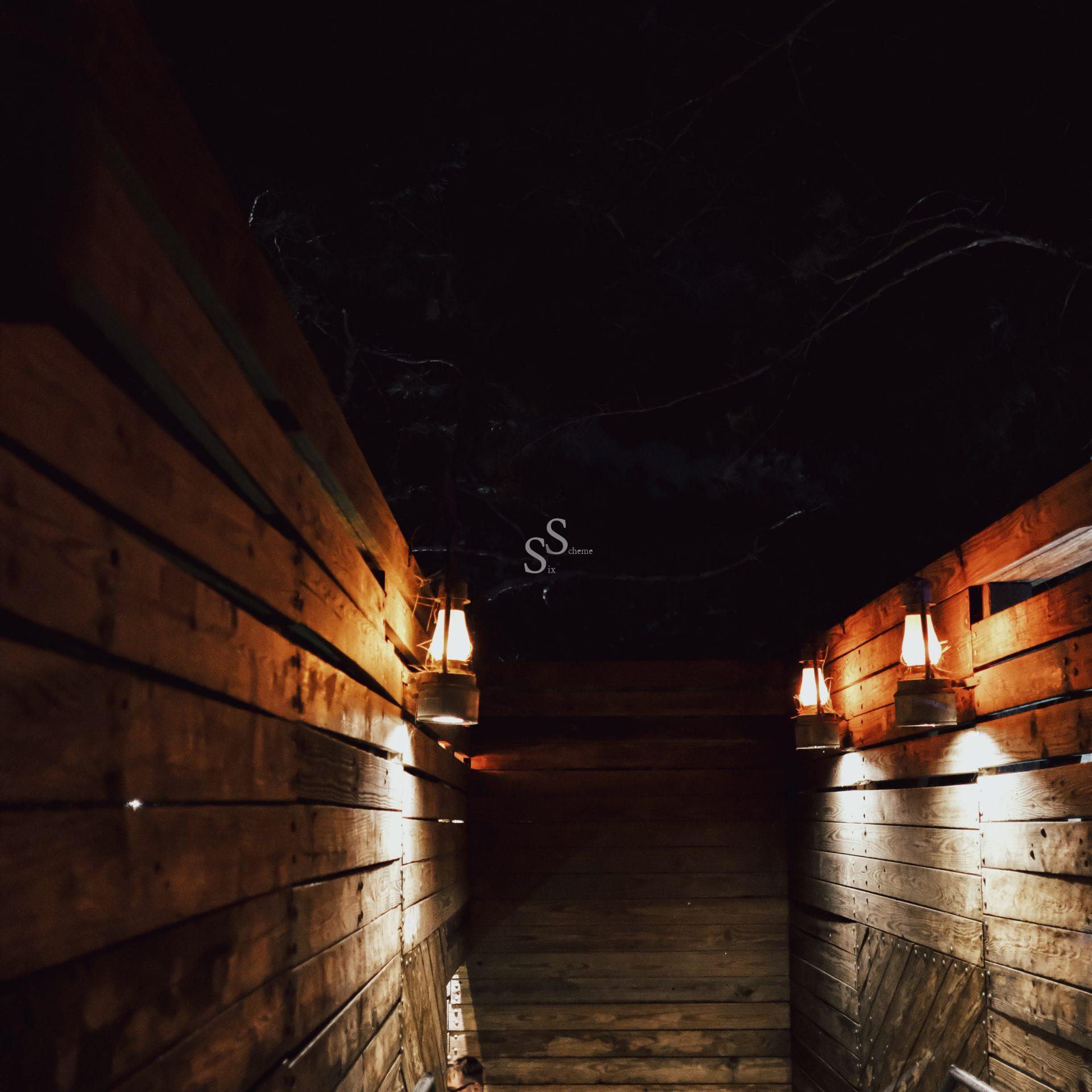 Dimly lit wooden corridor at night, illuminated by two lanterns mounted on the walls. The scene is rustic, with warm light casting shadows on the wood, and the sky above appears dark.