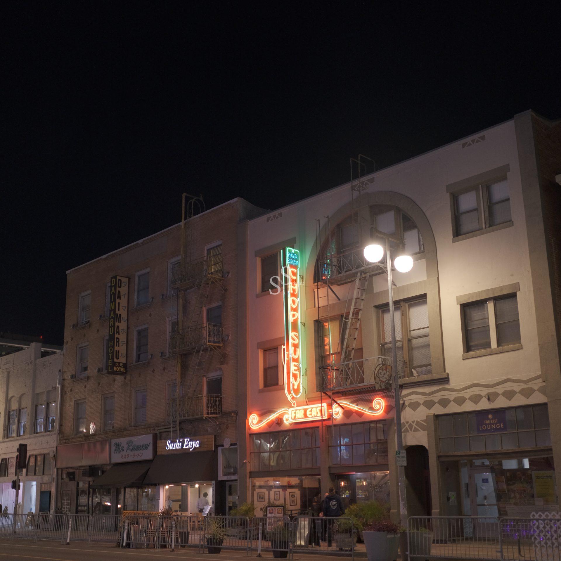 A nighttime street scene shows a row of buildings, with one having a neon sign reading, A Far East Chop Suey, above an entrance. The sidewalk is lit by street lamps, with shops and restaurants on the ground floor.